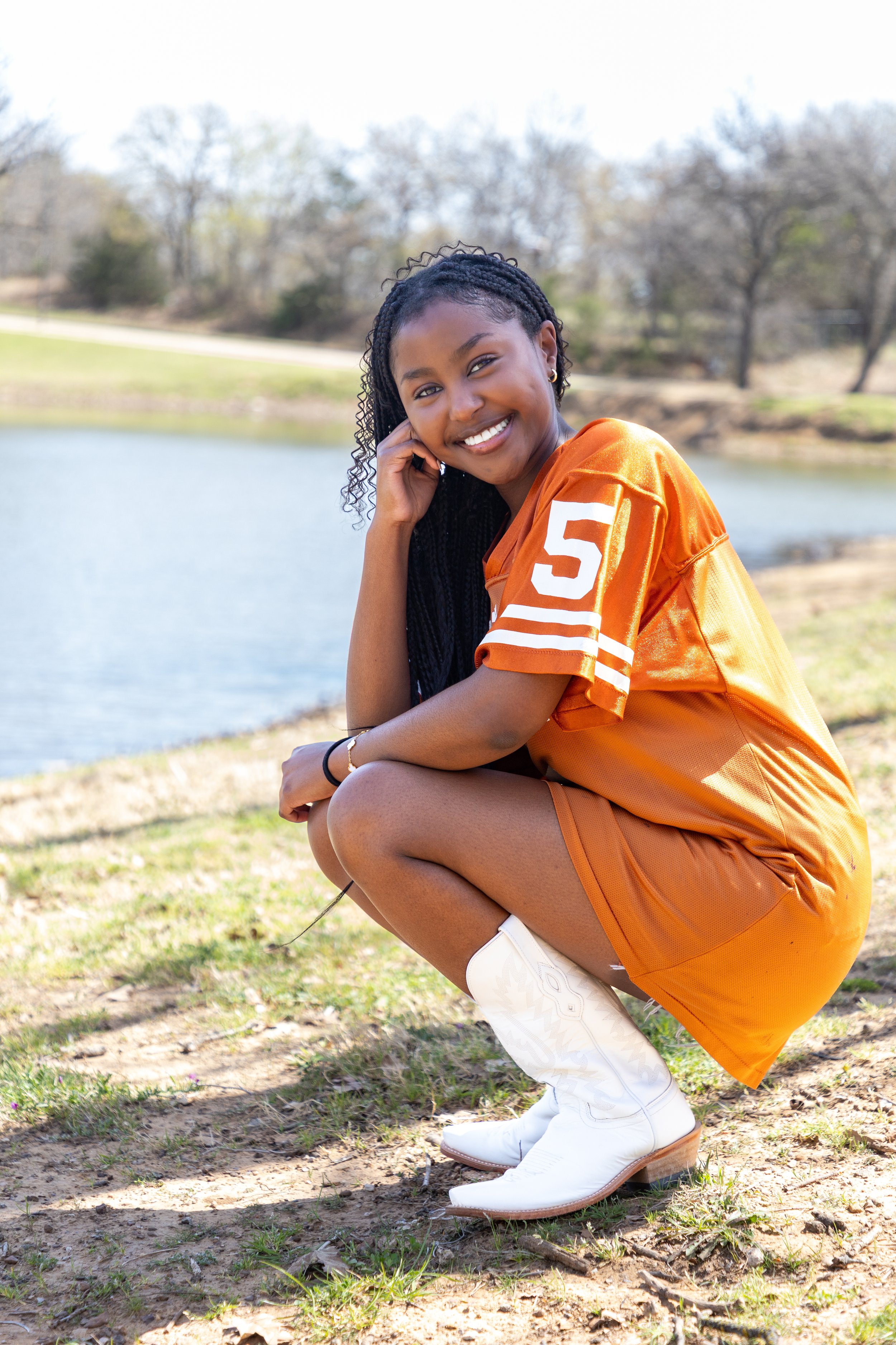A young woman crouching outdoors near a pond, smiling at the camera, wearing a football jersey with the number 5, orange shorts, white cowboy boots, and a black bracelet.