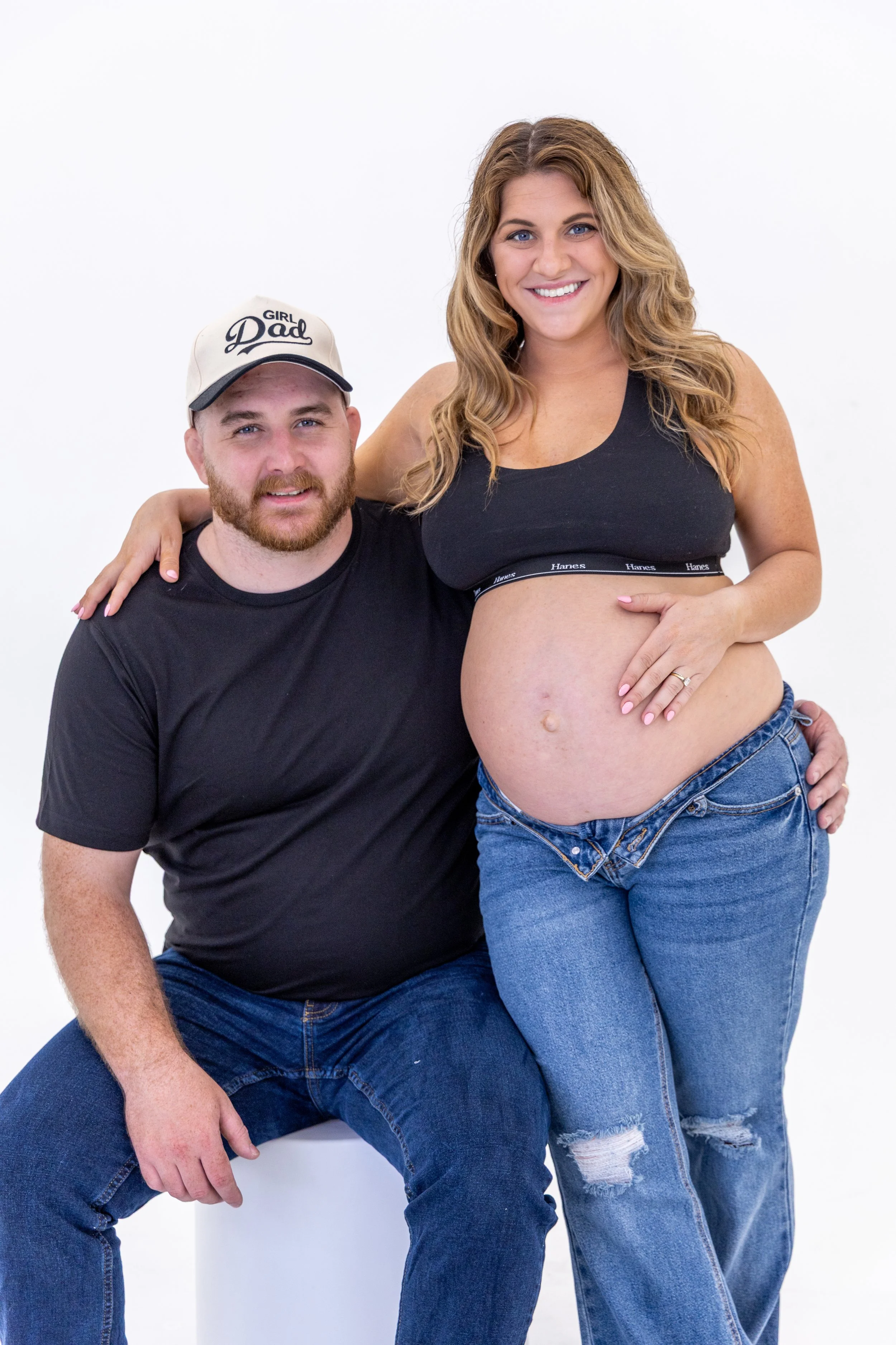 A couple, a man and a pregnant woman, sitting and standing together against a white background. The man is wearing a black t-shirt and a cap with "Girl Dad" printed on it. The woman is wearing a black sports bra with the brand name 'Hanes' and blue j