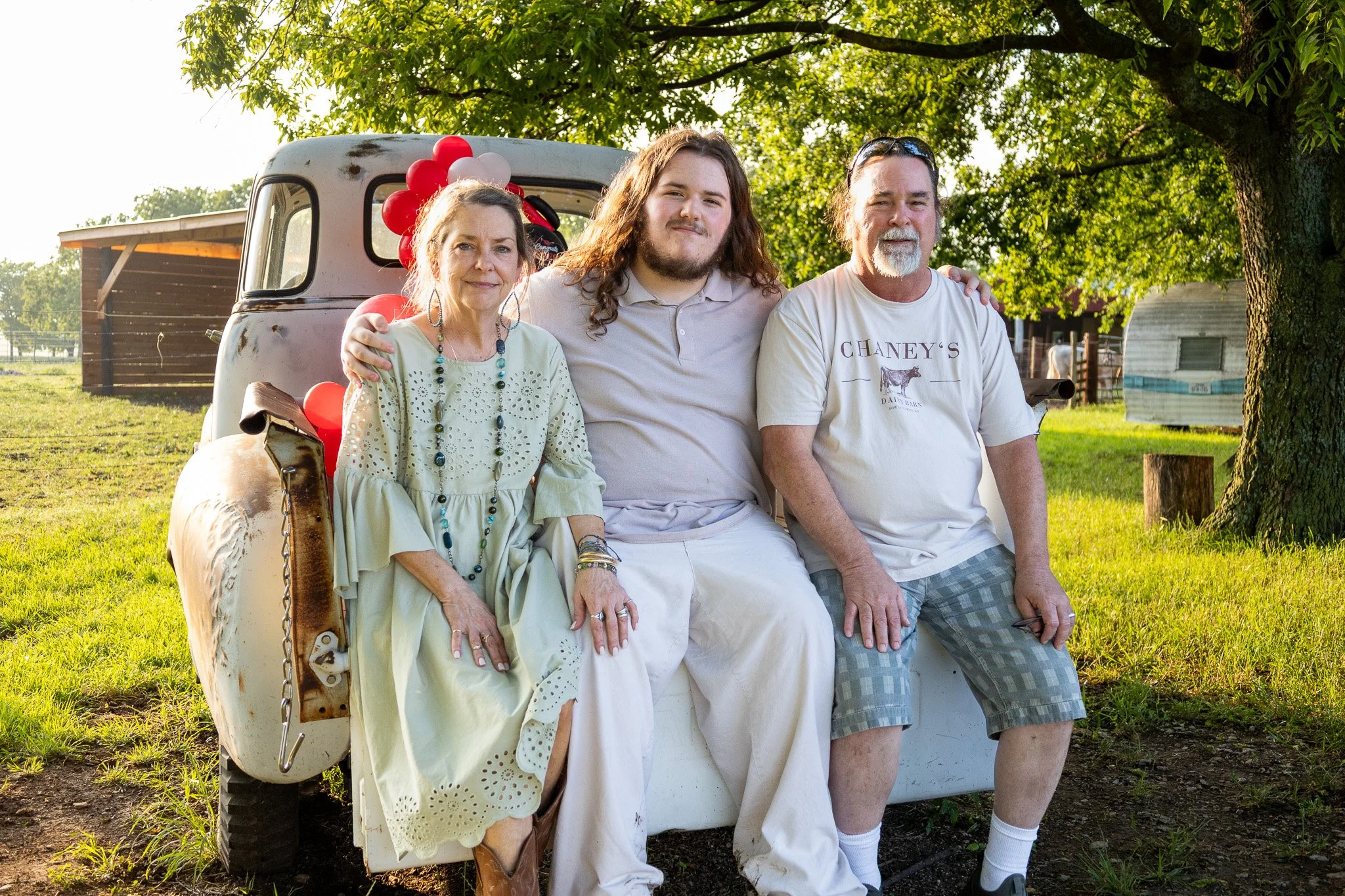 A family of three sitting on a vintage truck in a grassy area with trees in the background. It is daytime with sunlight shining through the leaves.