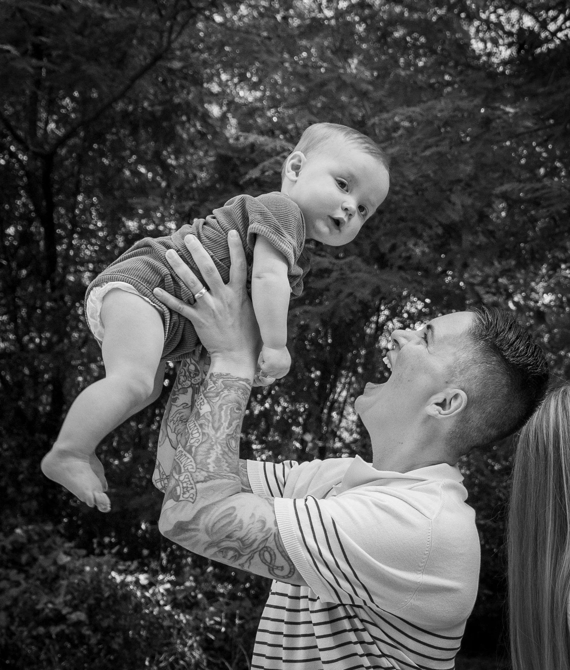 A man with tattoos on his arm and long hair holding a baby boy up in the air outdoors surrounded by trees, both smiling and looking at each other.
