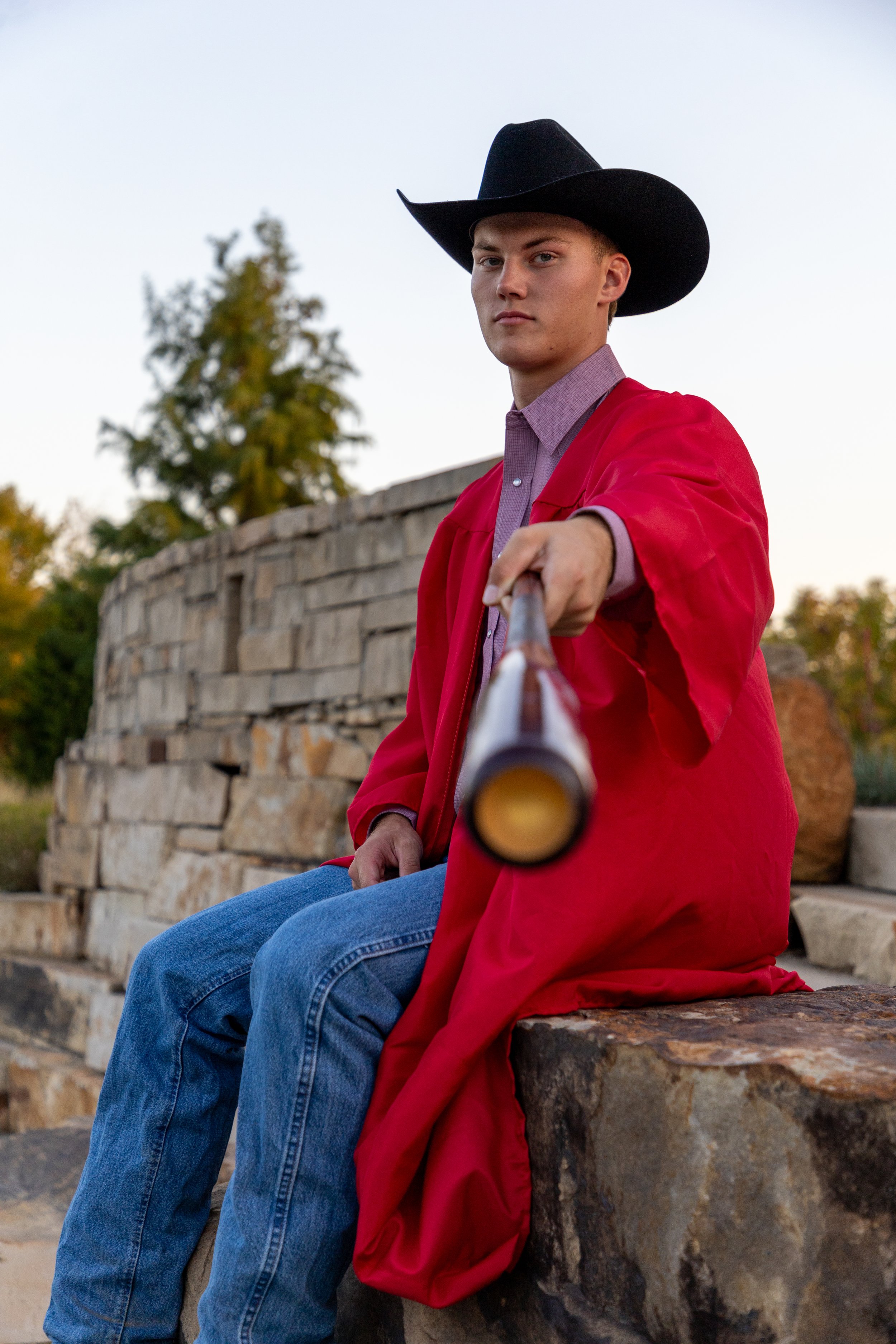 Young man wearing a black cowboy hat and a red coat, sitting on a stone ledge outside, pointing a rifle directly at the camera with a serious expression.