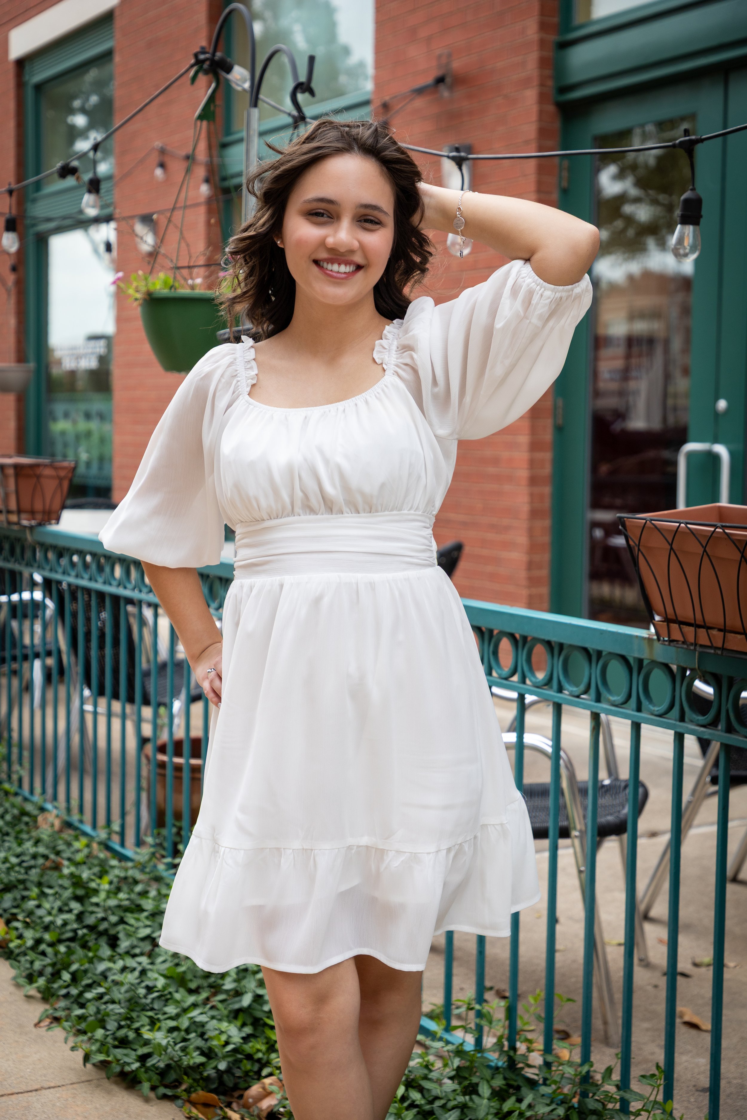 A young woman in a white dress smiling outdoors on a patio with brick walls, green window frames, and string lights.