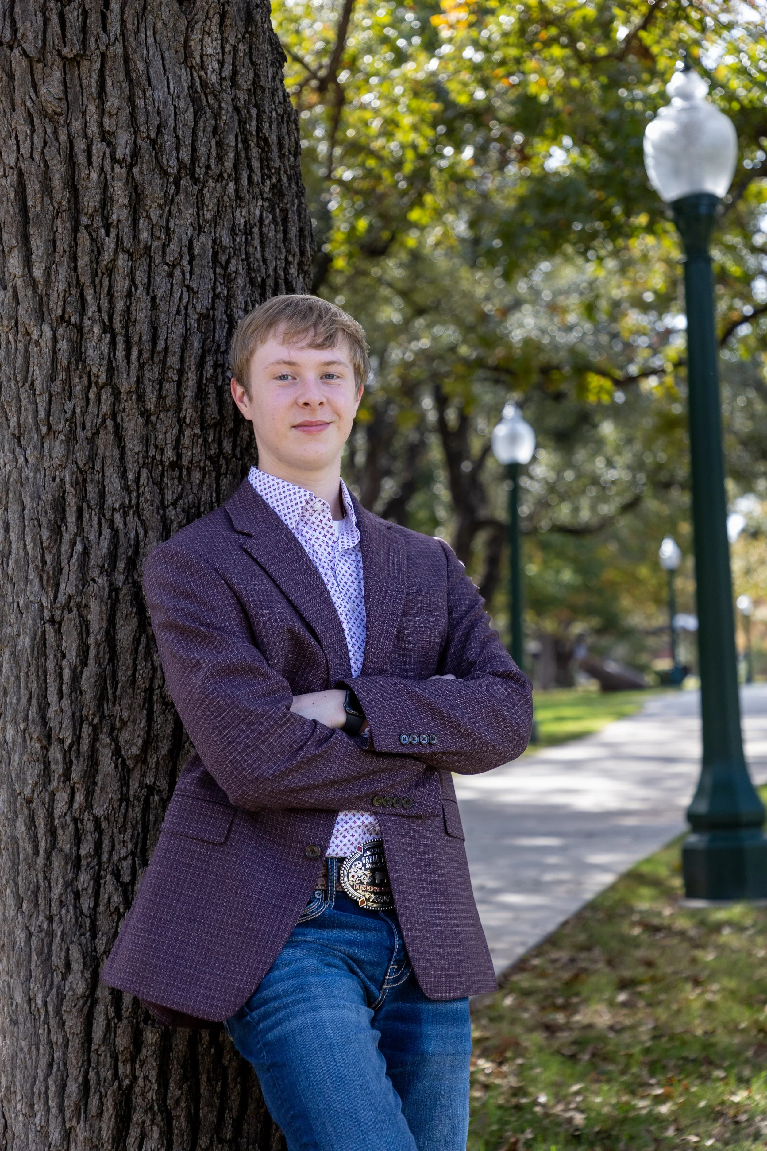A young man in a purple checkered blazer with arms crossed, leaning against a large tree in a park with a sidewalk and street lamps in the background.