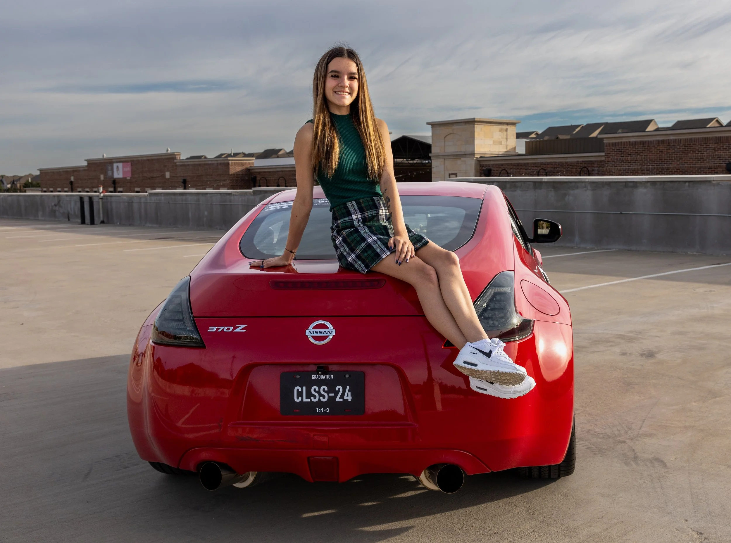 Young woman sitting on the trunk of a red Nissan 370Z sports car on top of a parking garage with buildings and a cloudy sky in the background.