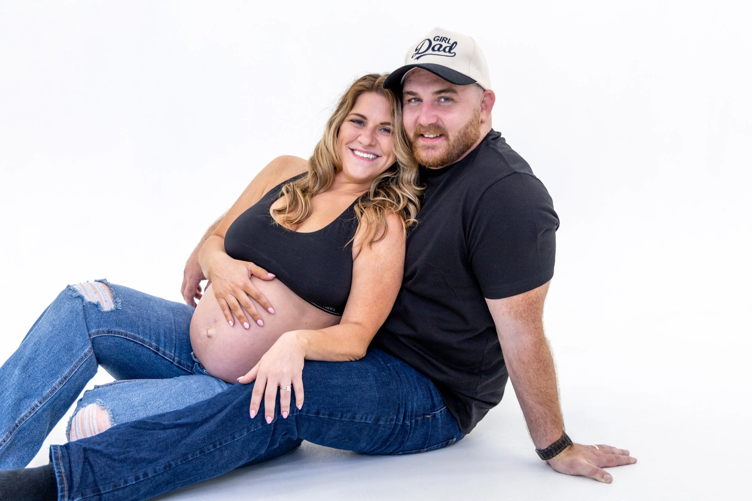 A pregnant woman and a man sitting together on the floor, smiling and embracing, against a plain white backdrop.