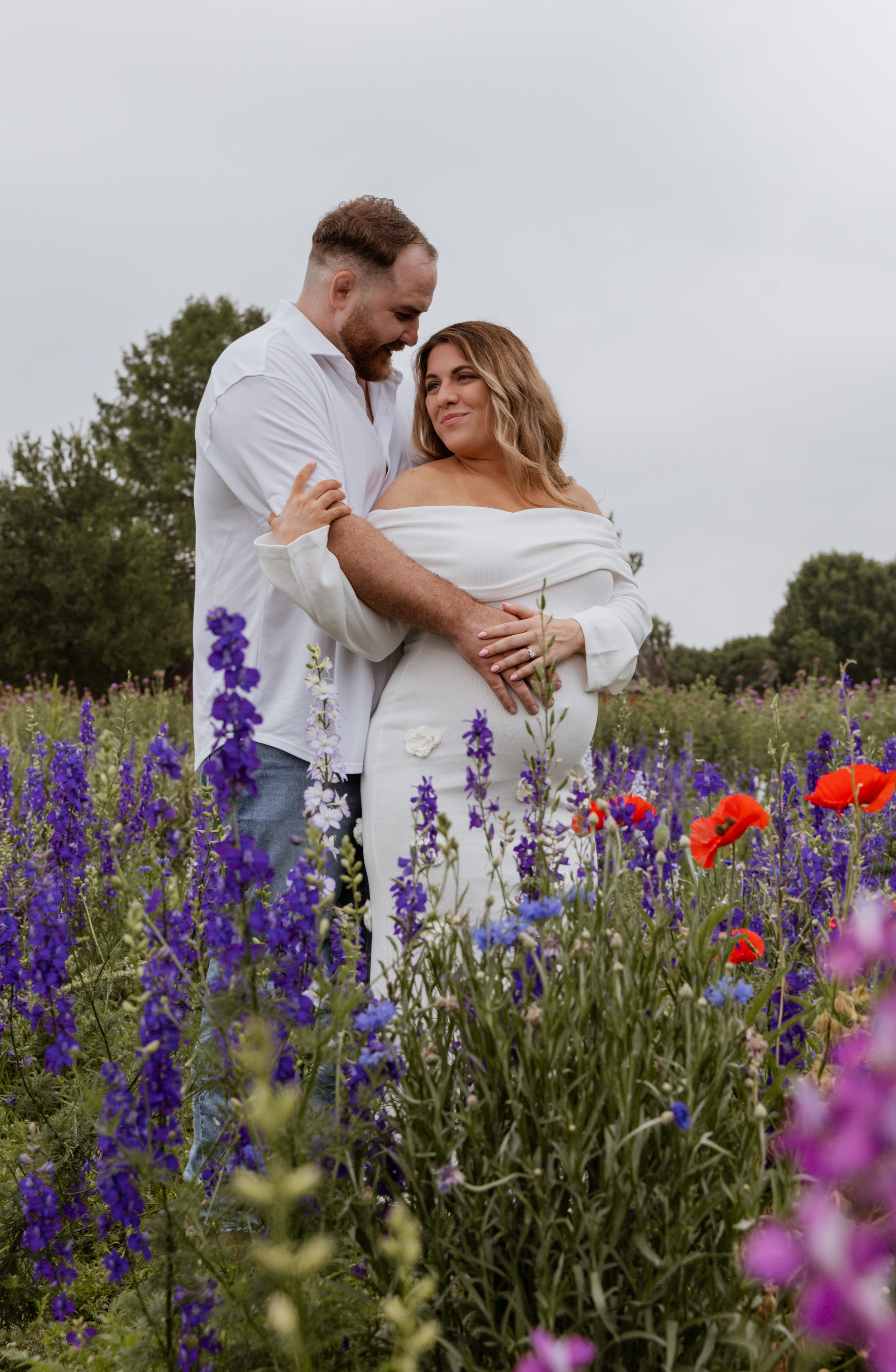 A couple standing in a field of purple and red flowers, with trees in the background, on an overcast day.
