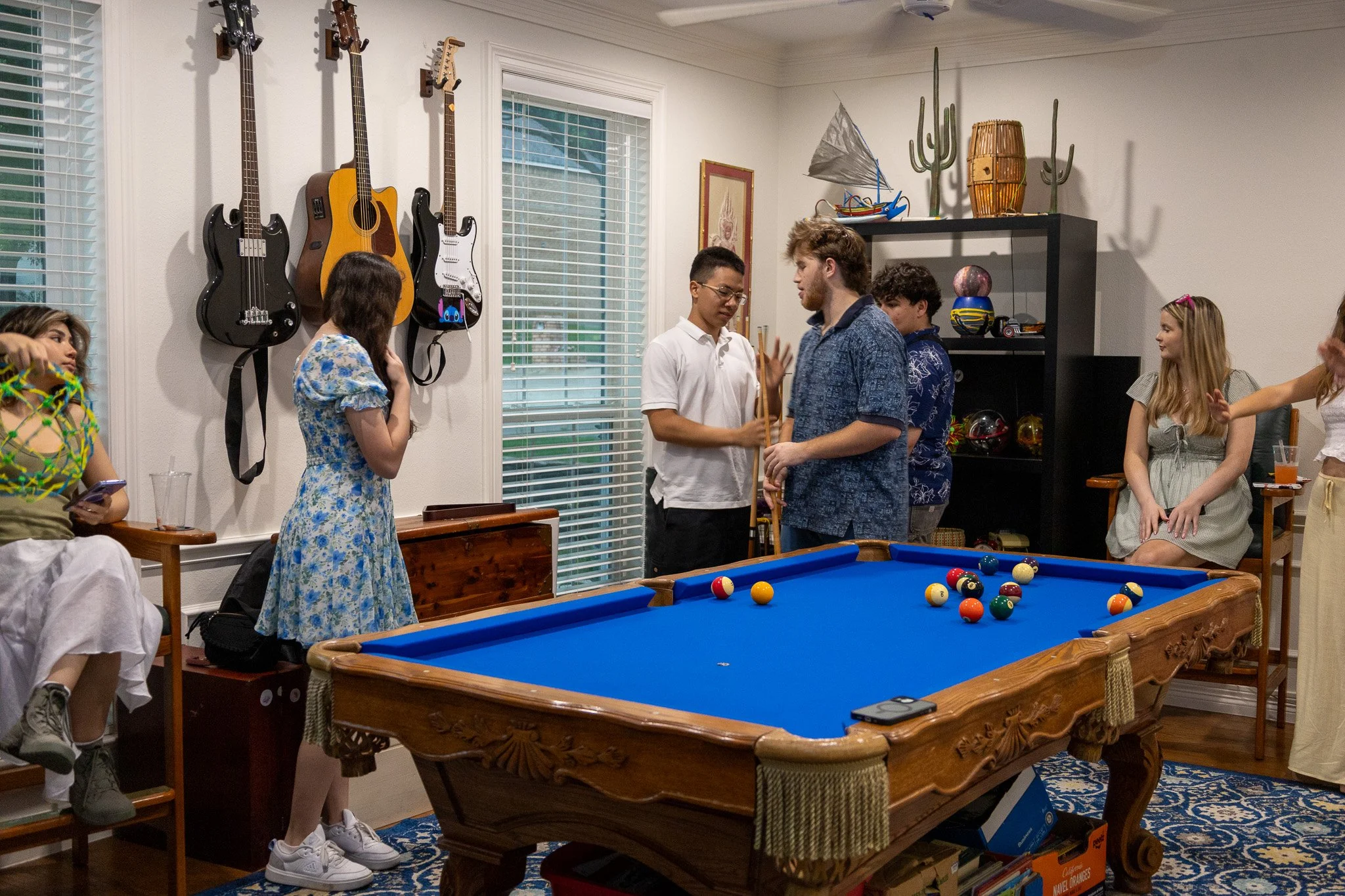 Group of young people socializing in a game room with billiards table, guitars on wall, and decorative items.