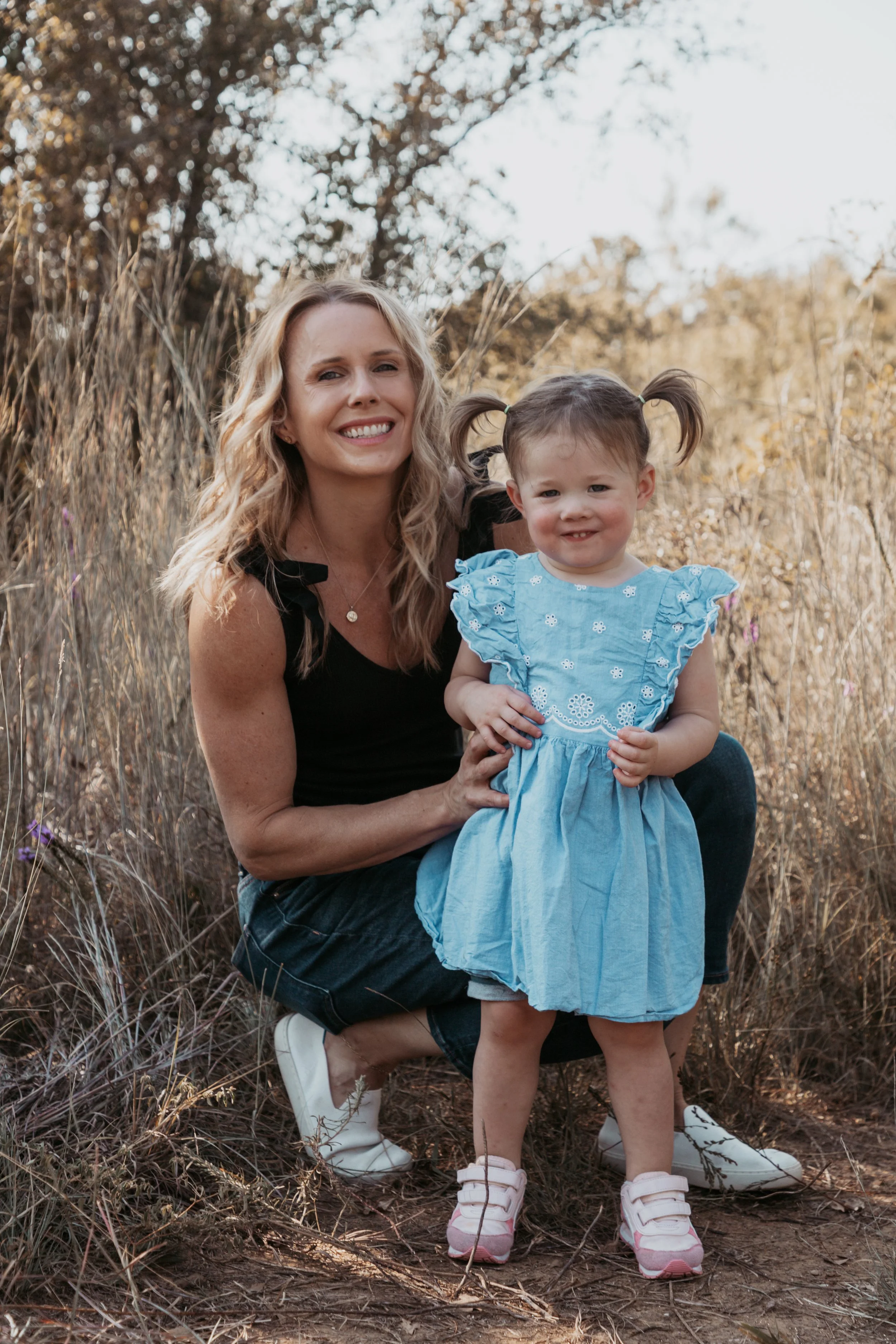 A woman kneeling outdoors in a natural setting with tall grasses and trees, smiling as she holds a young girl in a blue dress. They appear to be enjoying a moment together in a sunny, rural environment.