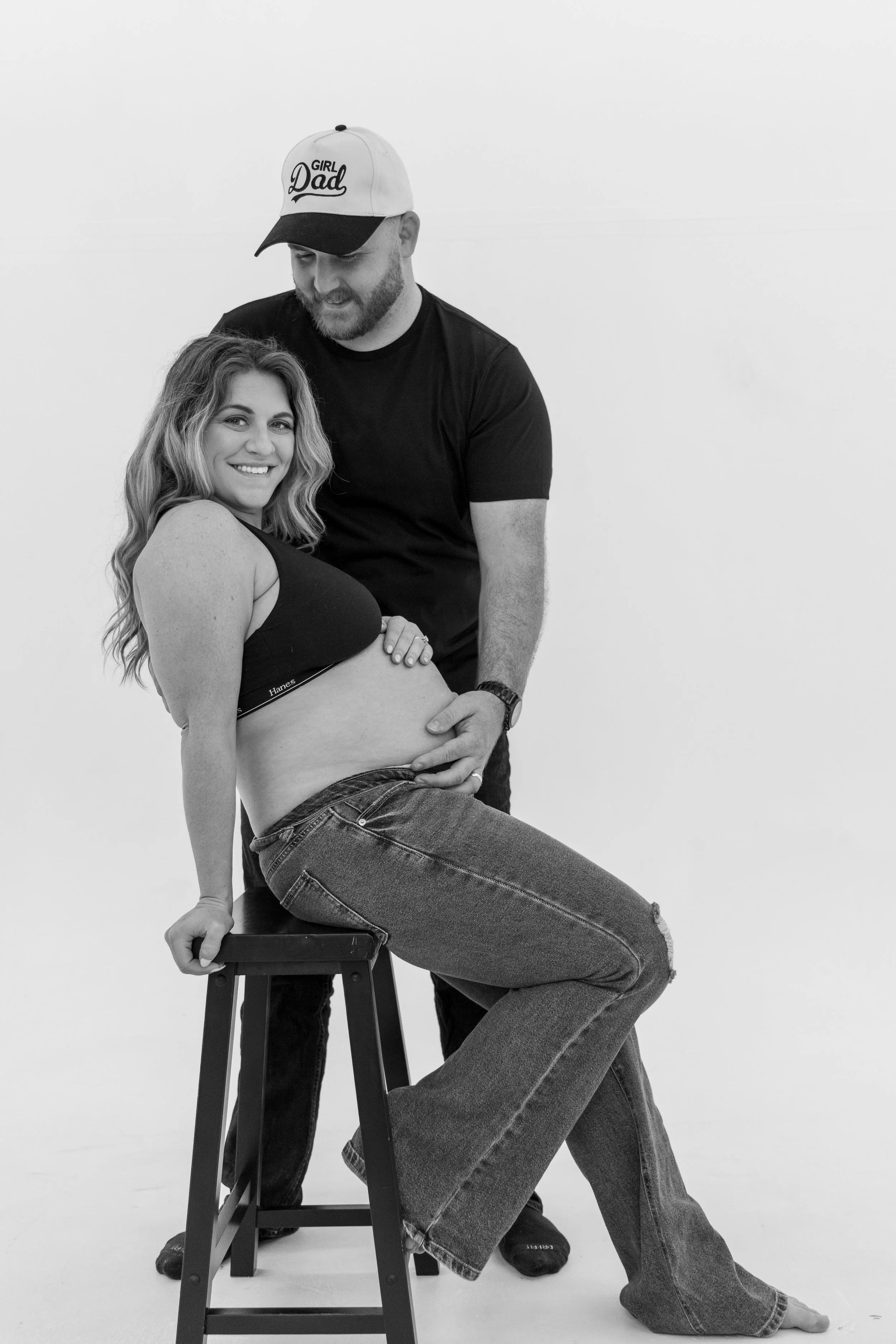 A pregnant woman sitting on a stool with her hand on her belly, smiling at the camera while a man stands behind her, looking down at her belly, both dressed casually, in front of a plain white background. The man is wearing a cap with the words "Girl