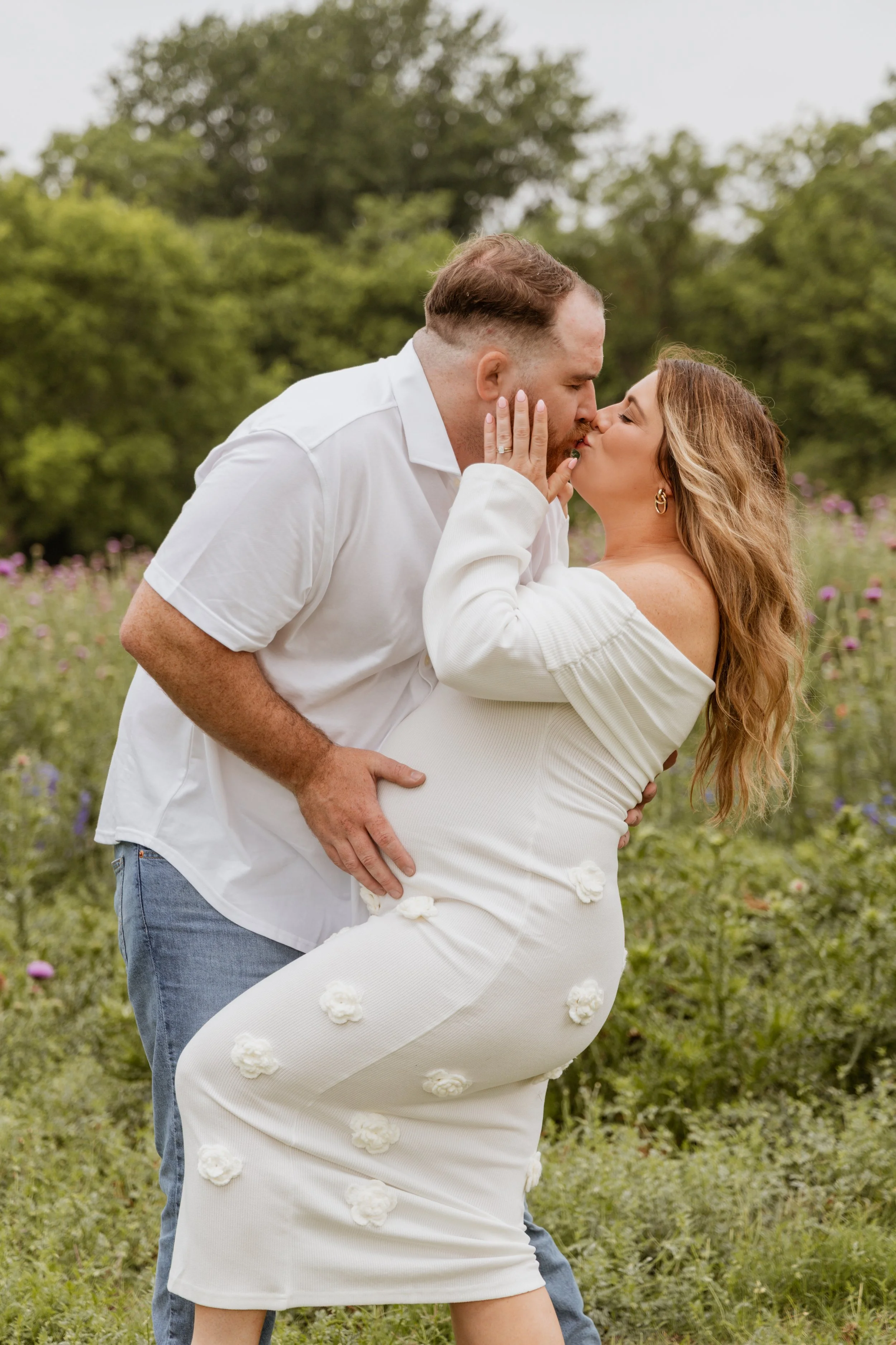 A couple kissing outdoors in a garden with greenery and flowers in the background.