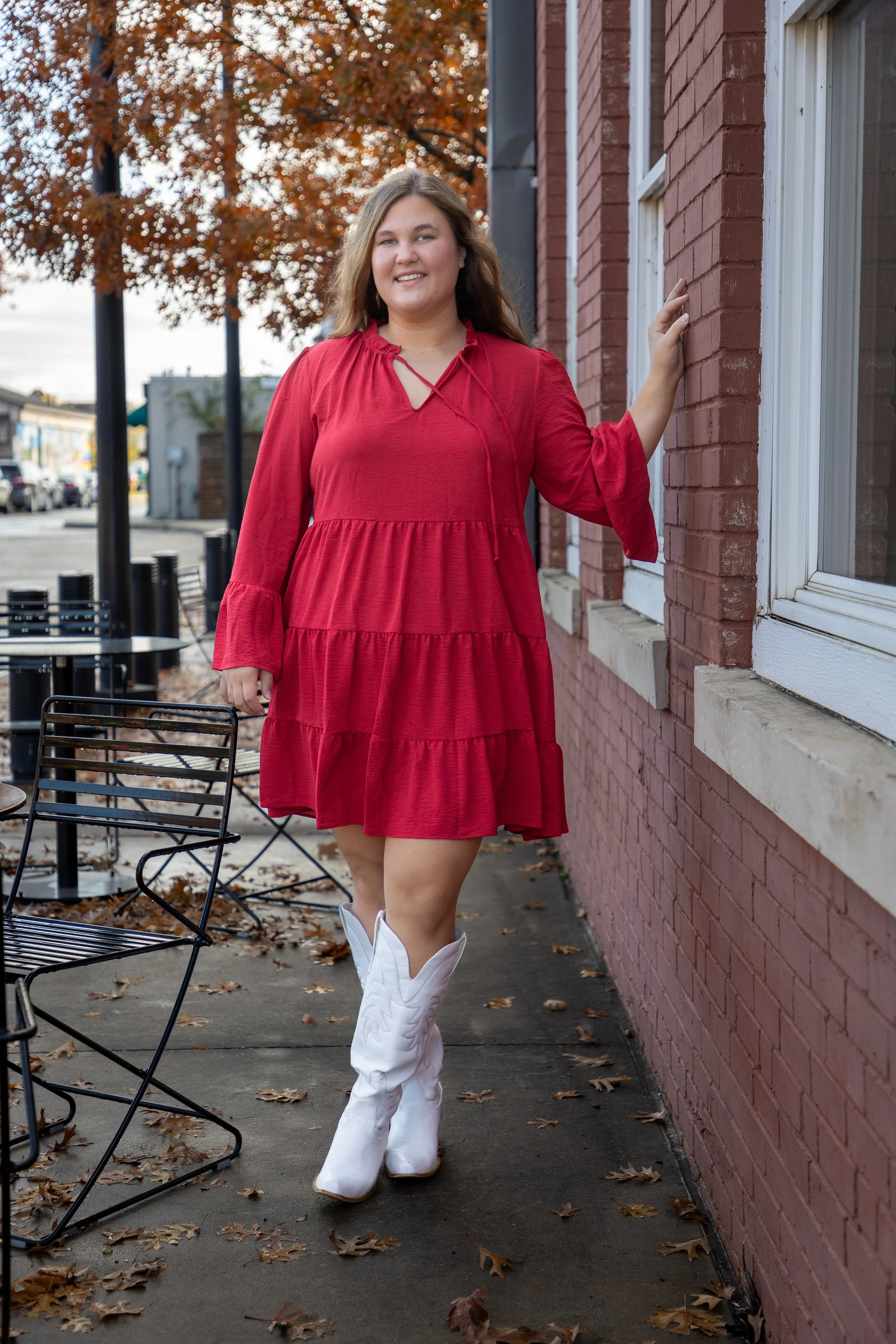 A woman with long brown hair, wearing a red dress and white cowboy boots, standing outside on a sidewalk next to a brick building, with autumn leaves on the ground and trees with orange leaves in the background.