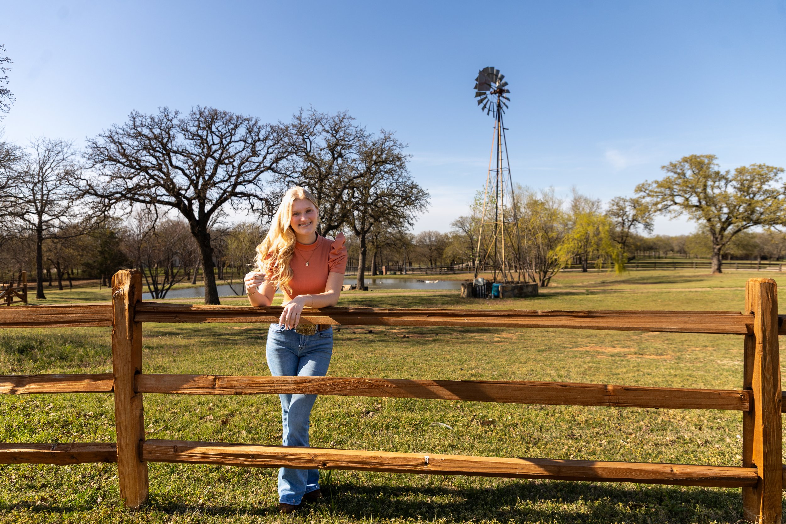 A young woman with long blonde hair wearing a peach-colored top and blue jeans standing behind a wooden fence in a park with trees, a pond, and a windmill in the background on a sunny day.