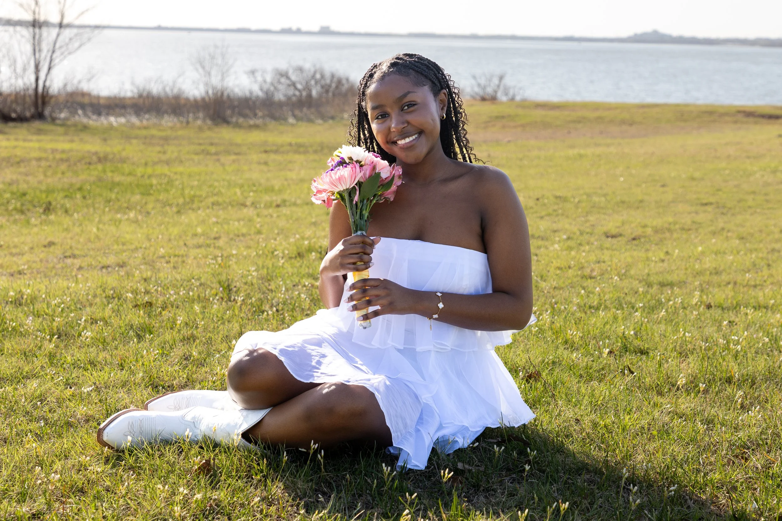 A young woman sitting on grass near a body of water, holding a bouquet of pink and white flowers, wearing a white strapless dress and white shoes, smiling at the camera.