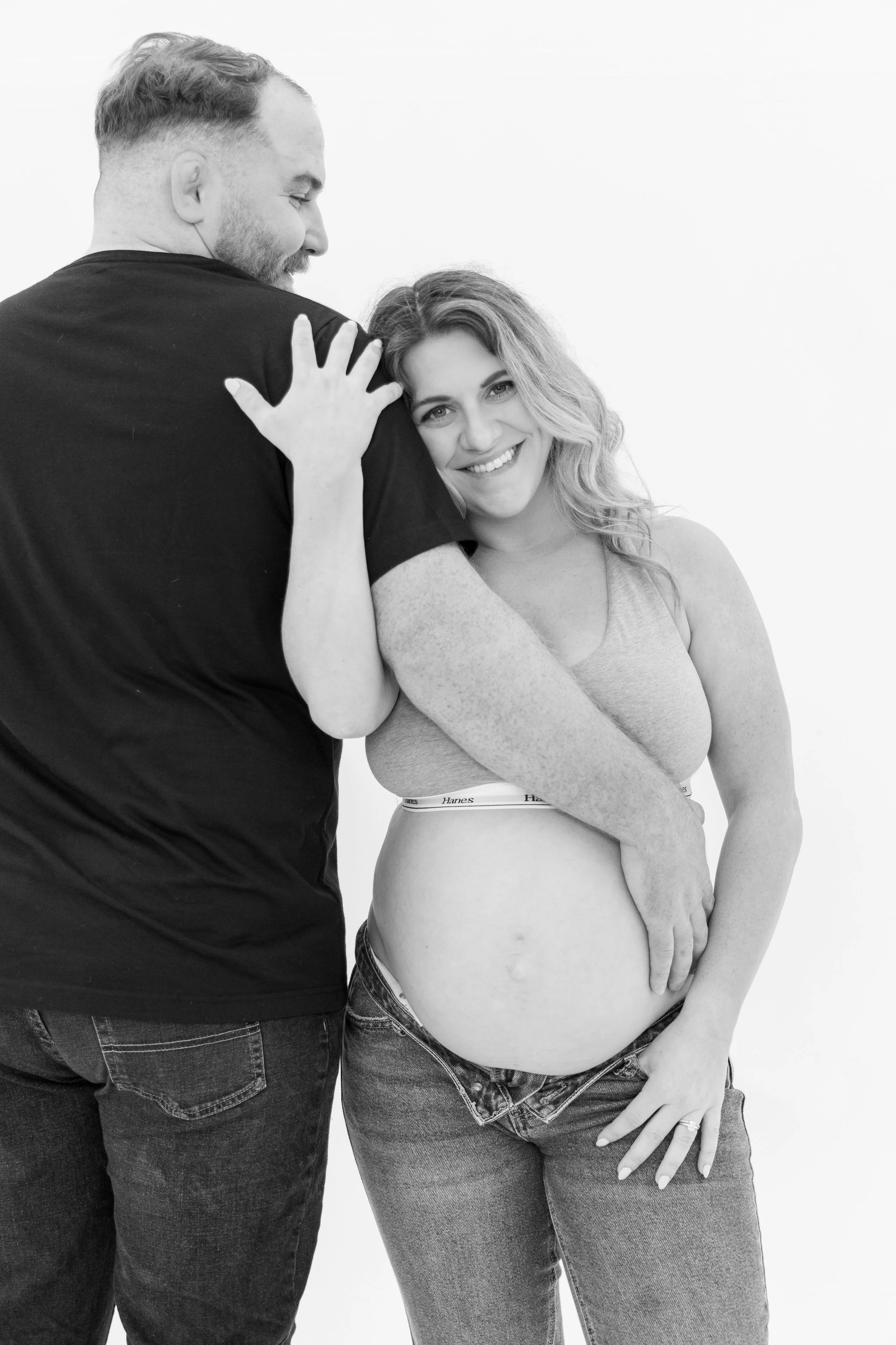A man and a pregnant woman share a joyful moment; the woman is smiling at the camera, and the man is gently touching her shoulder, both dressed casually, with the woman in jeans and a sports bra, and the man in a T-shirt, against a plain white backgr