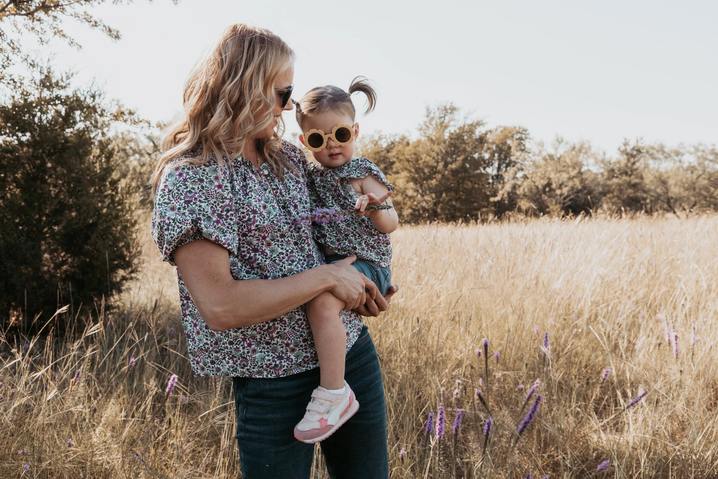 A woman with blonde wavy hair and sunglasses holding a young girl with sunglasses and a ponytail, standing in a grassy field with trees in the background.