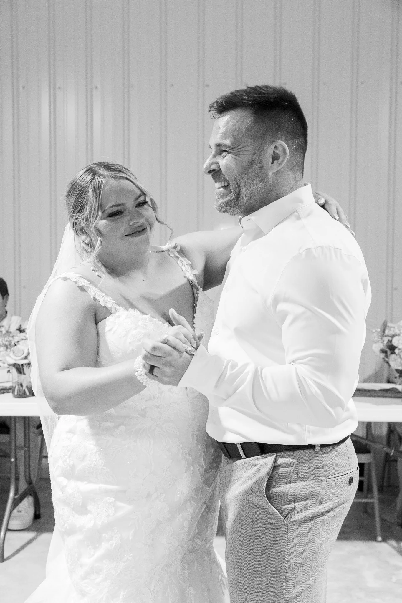 A black and white photo of a bride and groom sharing a dance, smiling and gazing at each other, at their wedding reception.