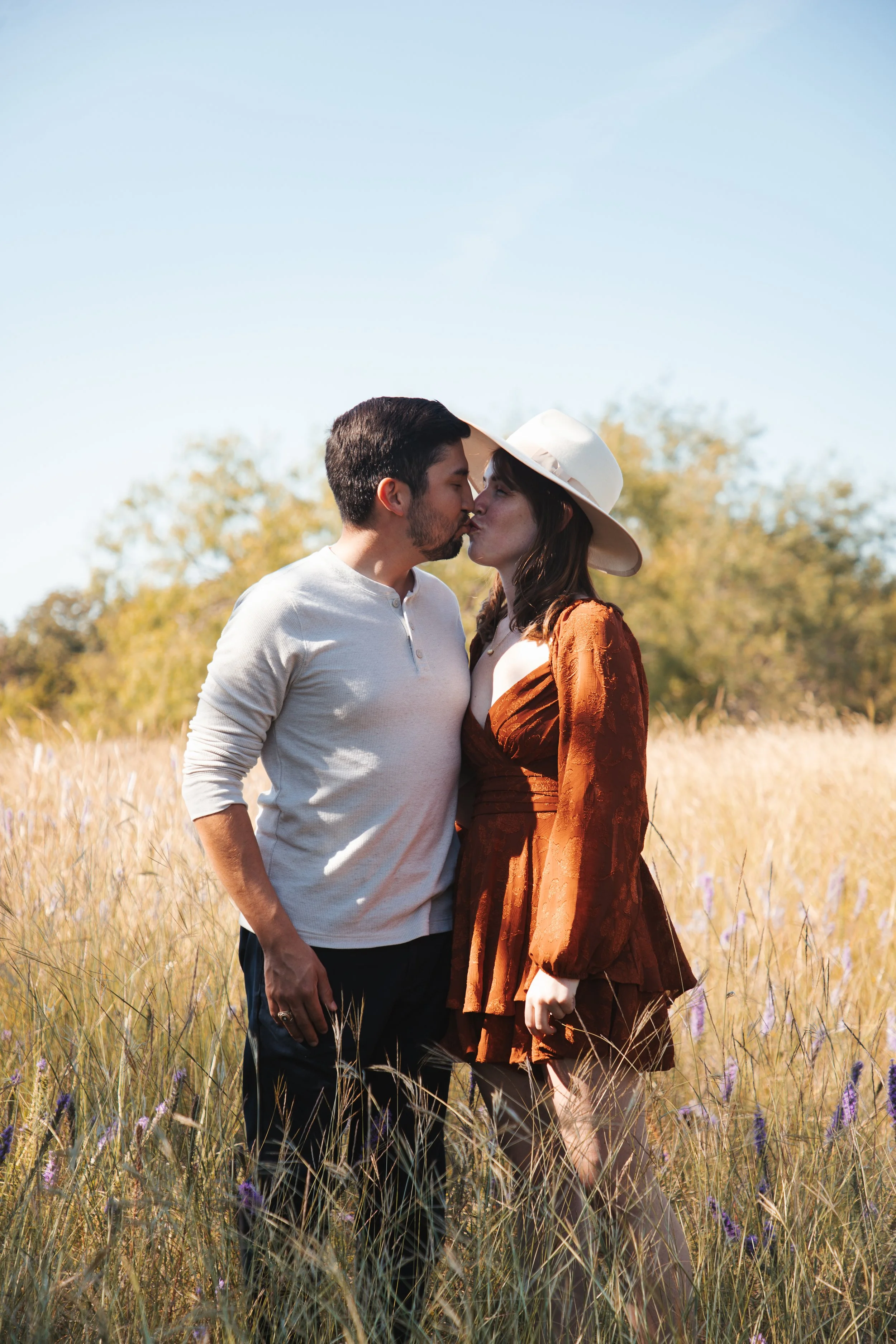 A couple sharing a kiss in a grassy field during daytime, with trees and a clear blue sky in the background.