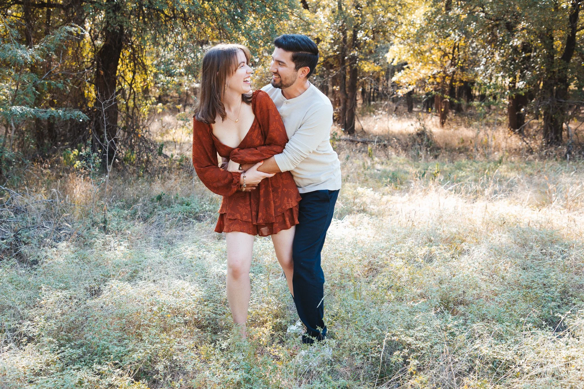 A young couple laughing and holding each other in a sunny forested area during fall.