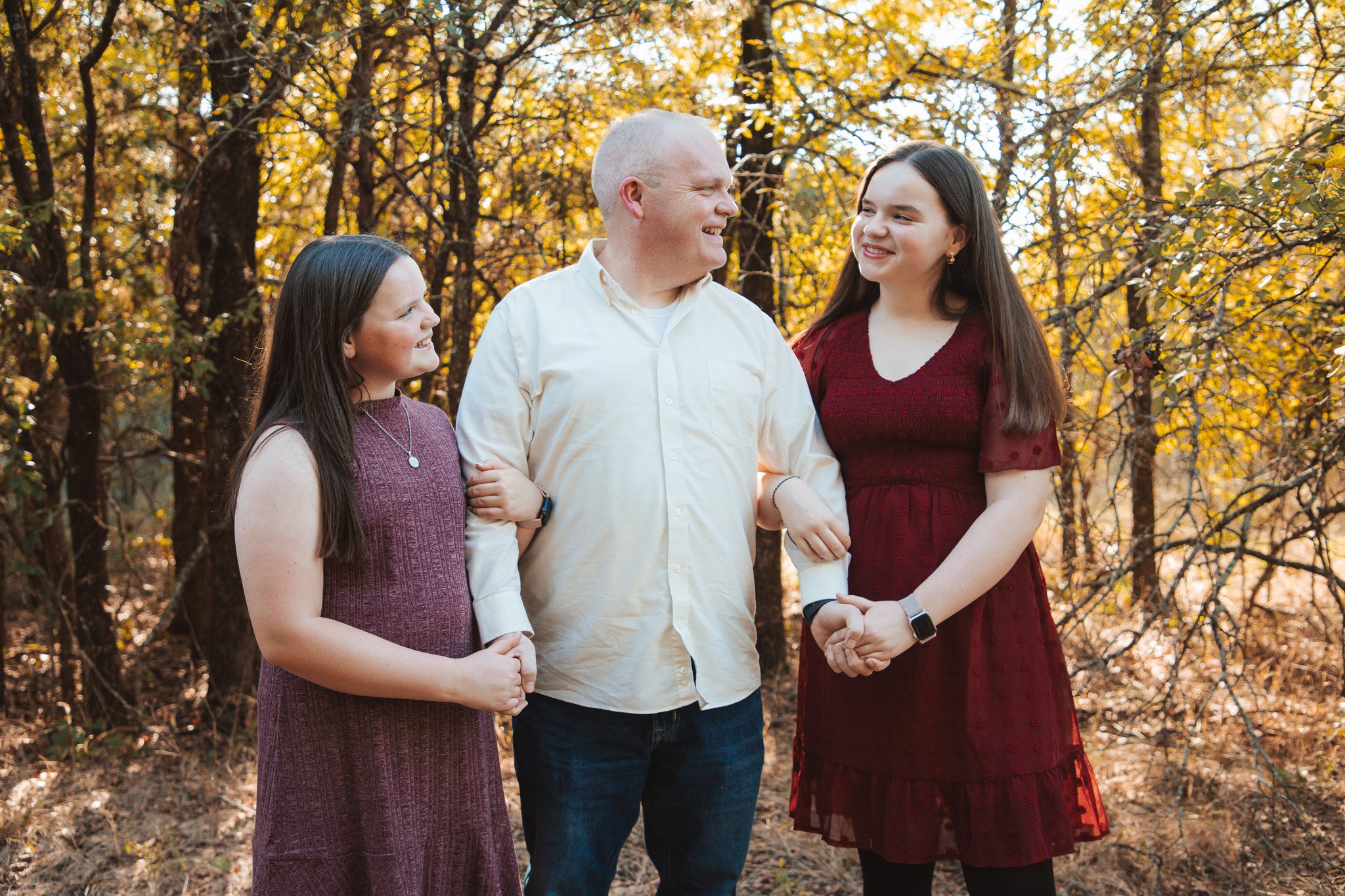 A family of four, including a man, woman, and two girls, standing outdoors in a wooded area with autumn foliage. They are smiling and looking at each other.