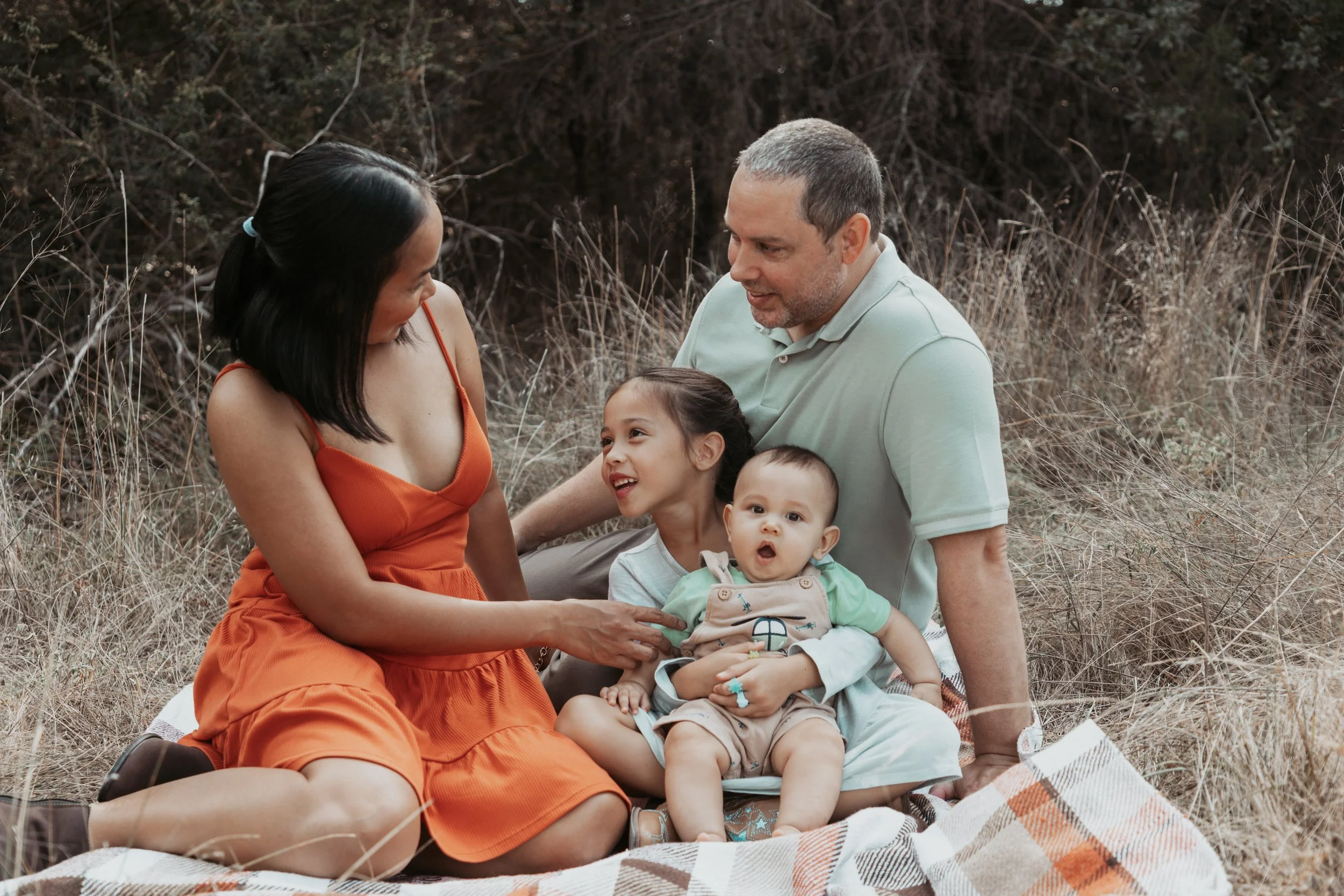 A family of four enjoying time outdoors on a picnic blanket in a grassy area with trees in the background, smiling and interacting with each other.
