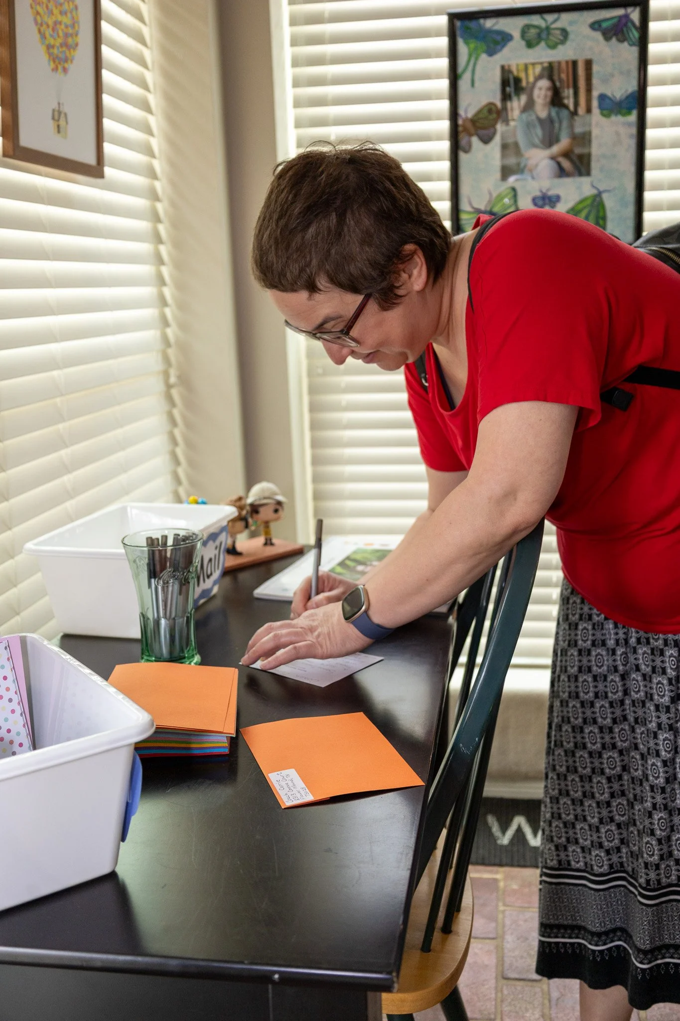 Woman in red shirt writing on a piece of paper at a black dining table with colorful index cards and storage bins, near window blinds and framed photos on the wall.