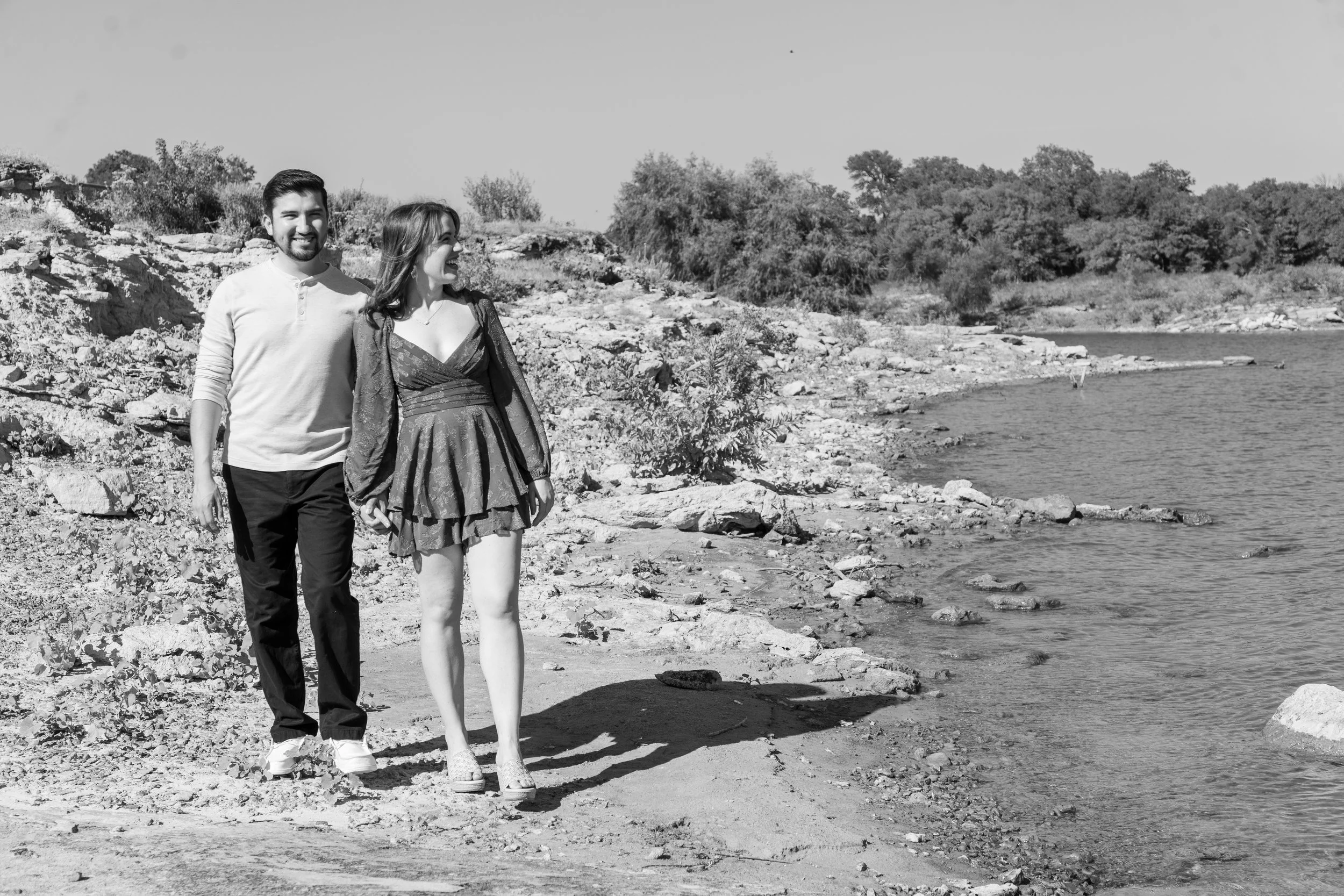 A man and woman walking along a rocky shoreline with trees in the background.