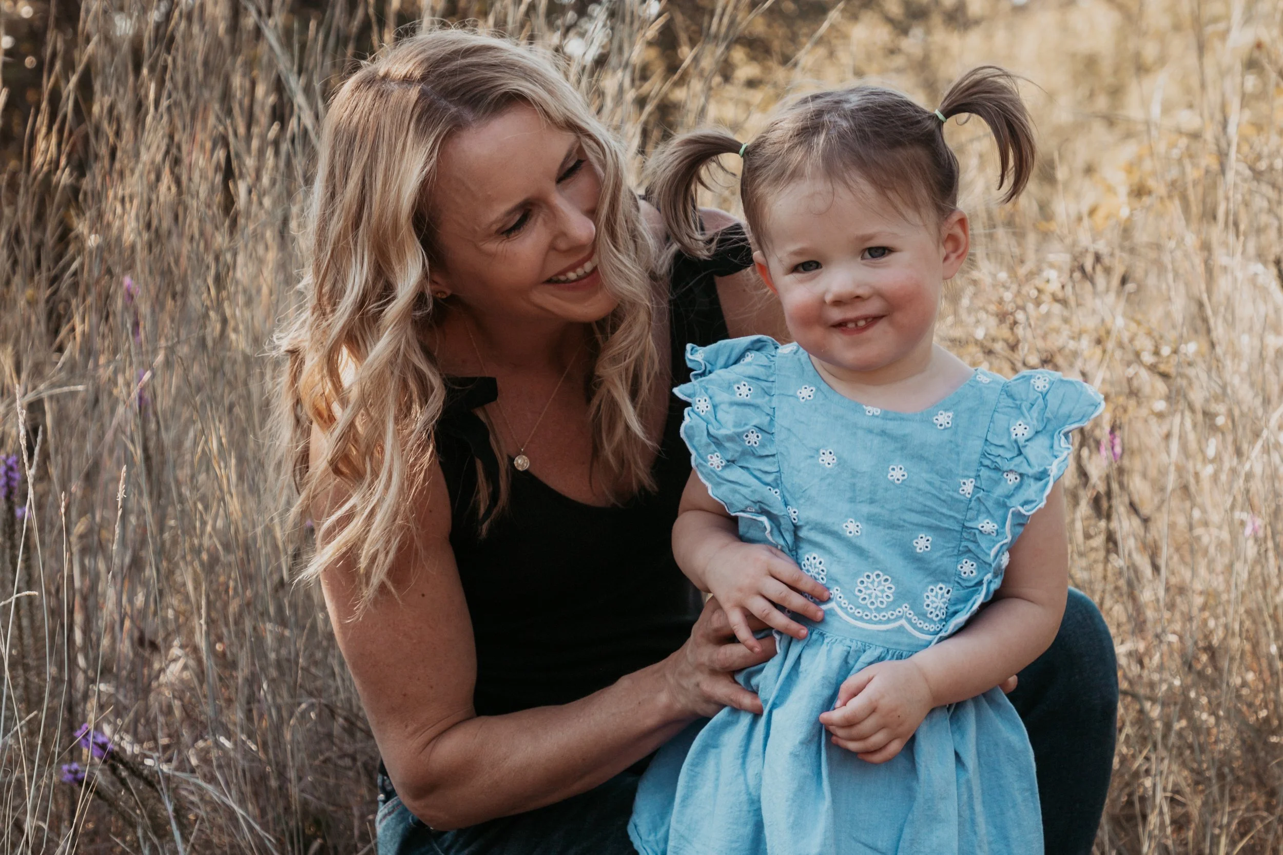 A woman with long blonde hair and a girl with pigtails wearing a blue dress look at the camera in a field of tall golden grass.