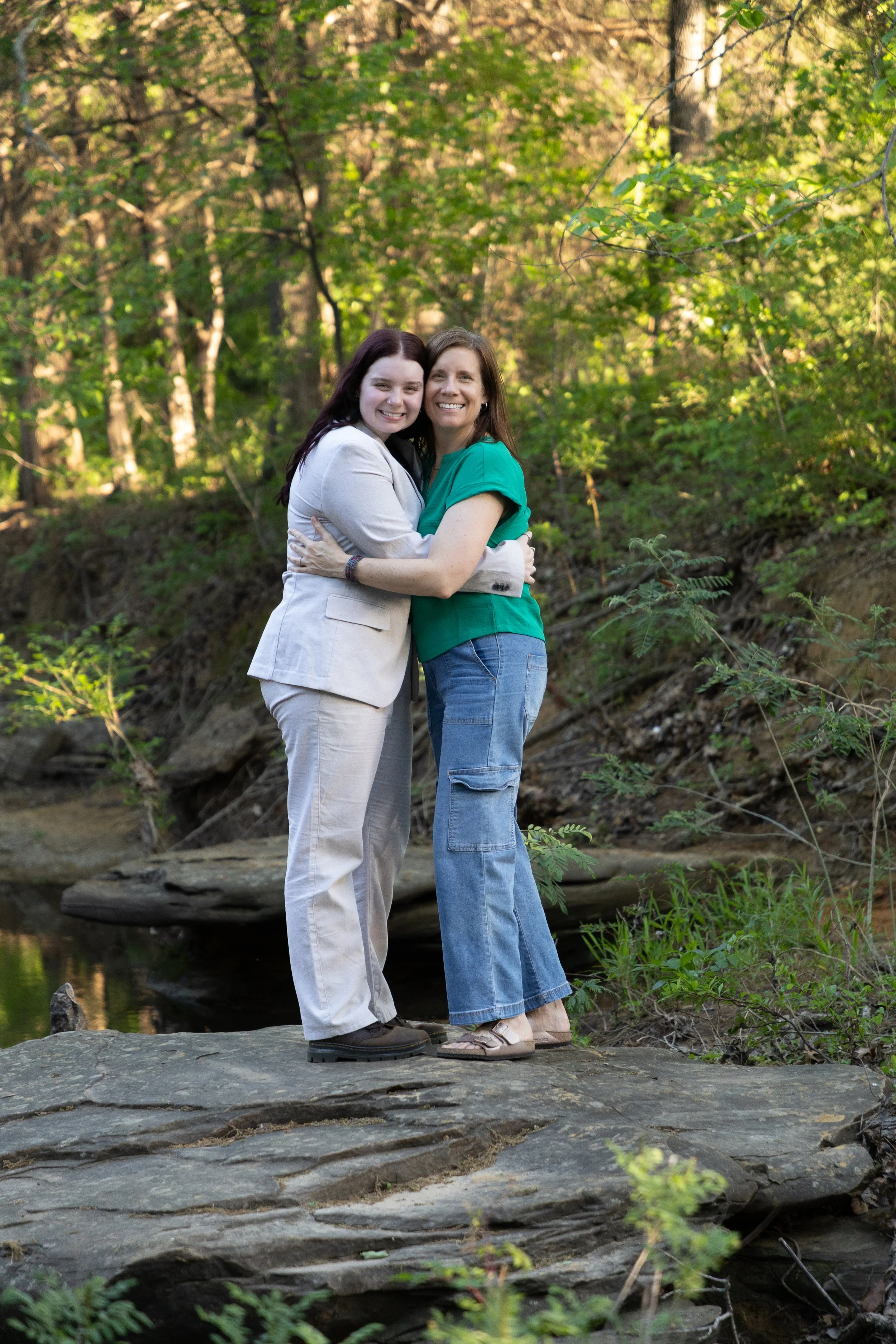 Two women hugging each other outdoors in a forested area near a small stream, smiling at the camera.
