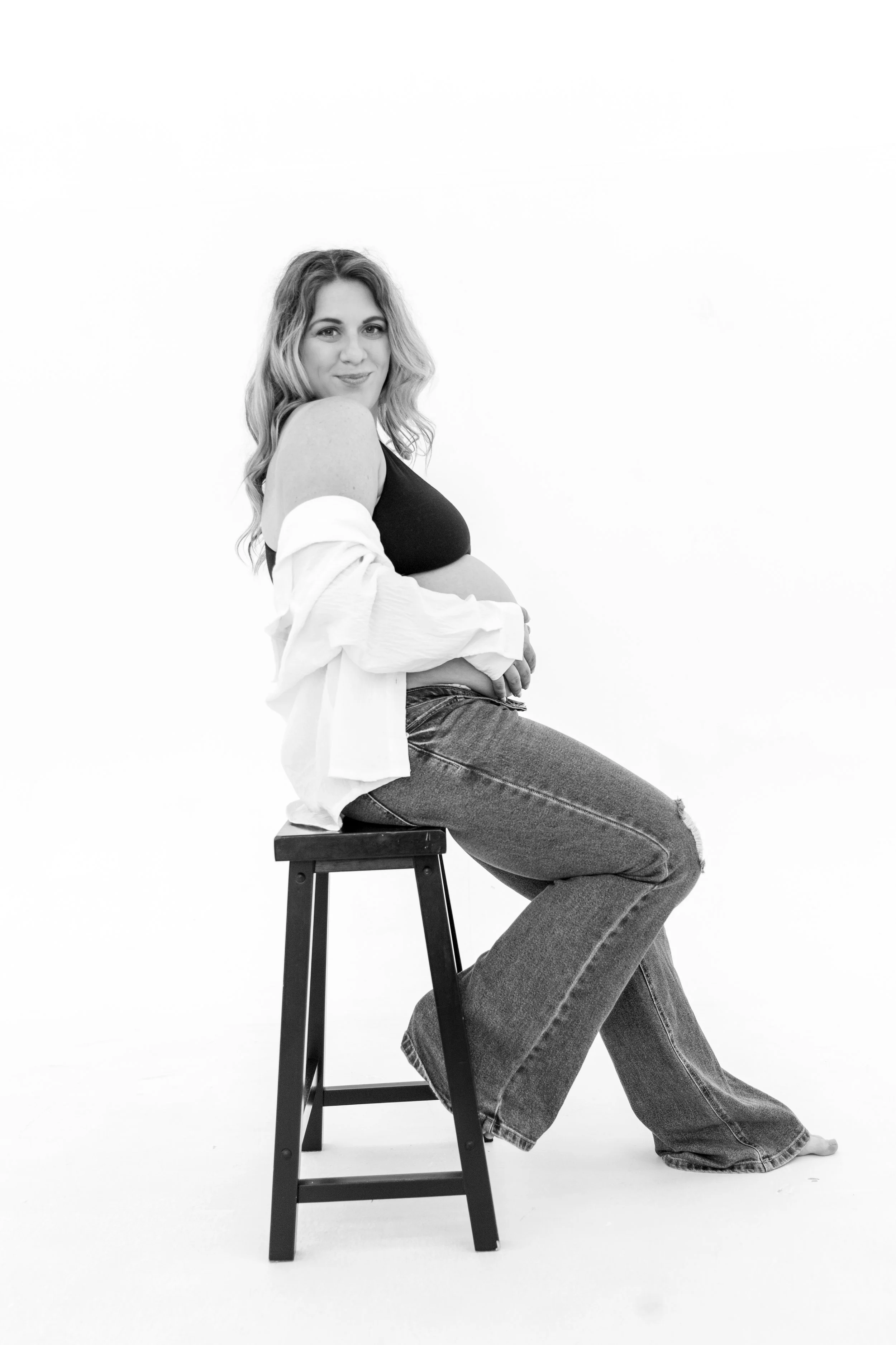 Black and white photo of a pregnant woman sitting on a stool, facing the camera, with one knee on the ground, dressed in jeans and a tank top, smiling.