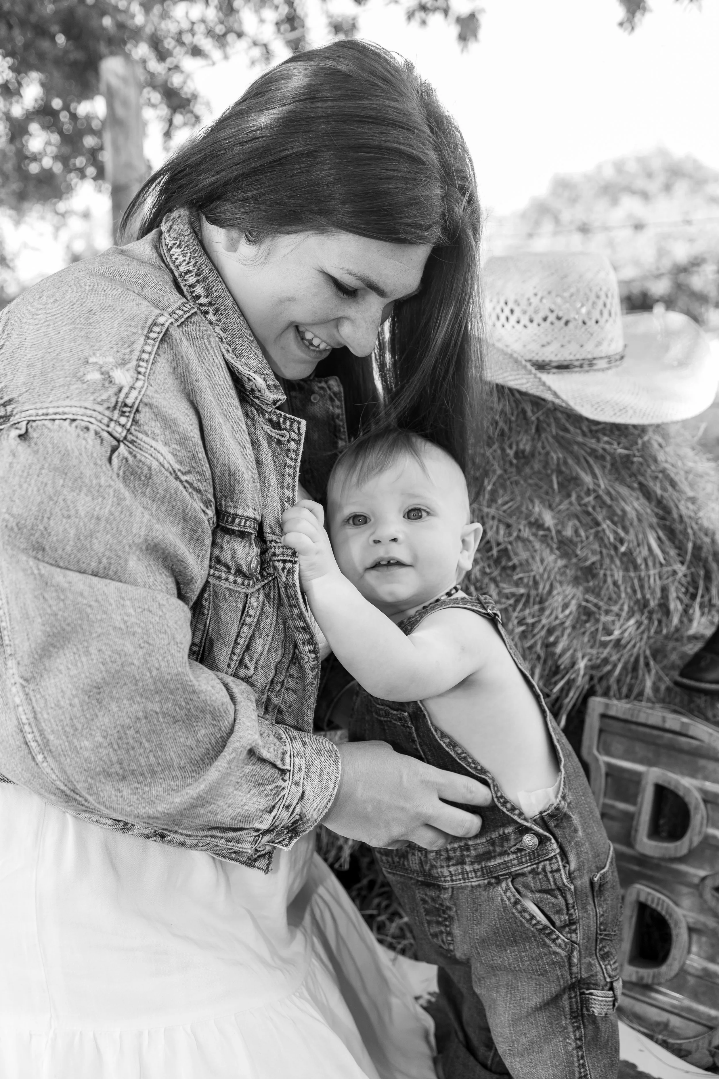 A woman and a baby in a rural outdoor setting, with hay bales and a straw hat in the background, captured in black and white.