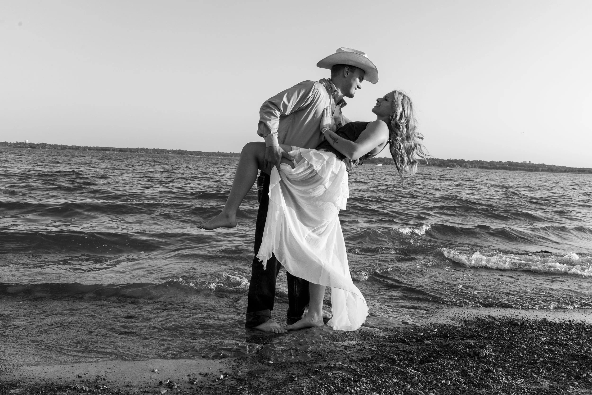 A couple stands in shallow water at the beach, with the man wearing a cowboy hat and the woman smiling as he holds her in his arms, both smiling and looking at each other.