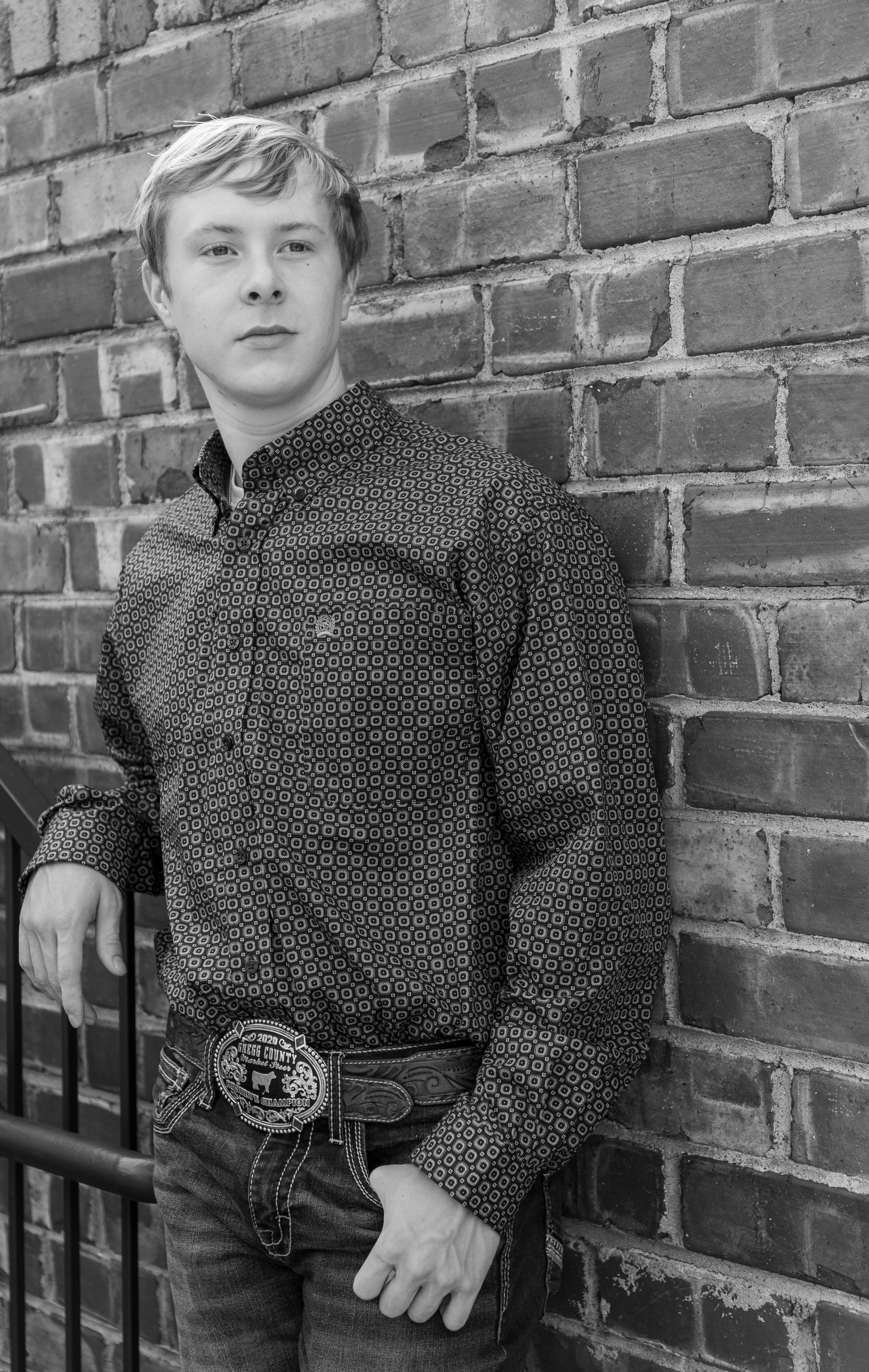 A young man with light hair, wearing a patterned button-up shirt and jeans, standing against a brick wall, posing with one hand on a railing.