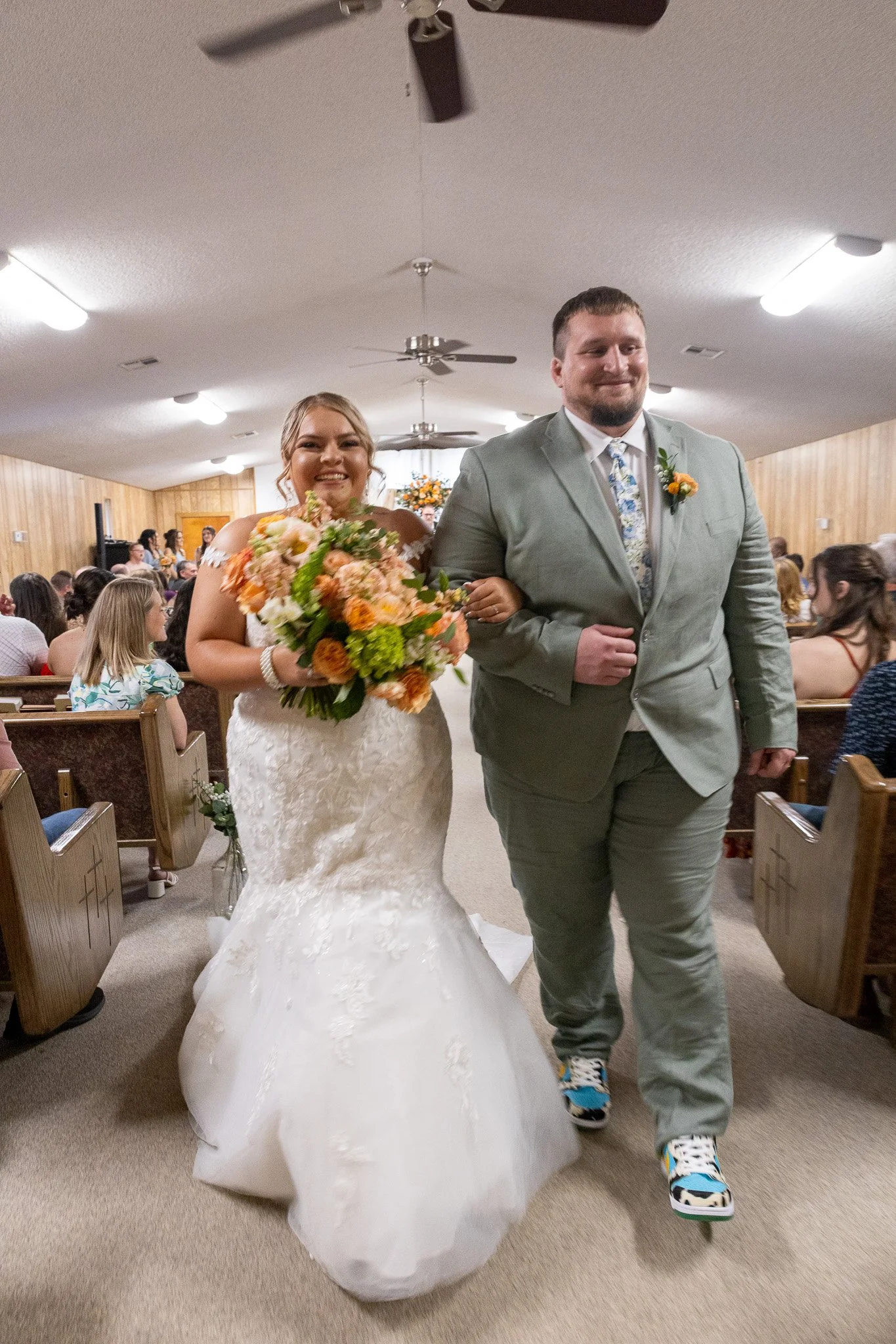A bride and groom walking down the aisle at their wedding ceremony, with guests seated on either side and wooden walls in the background.