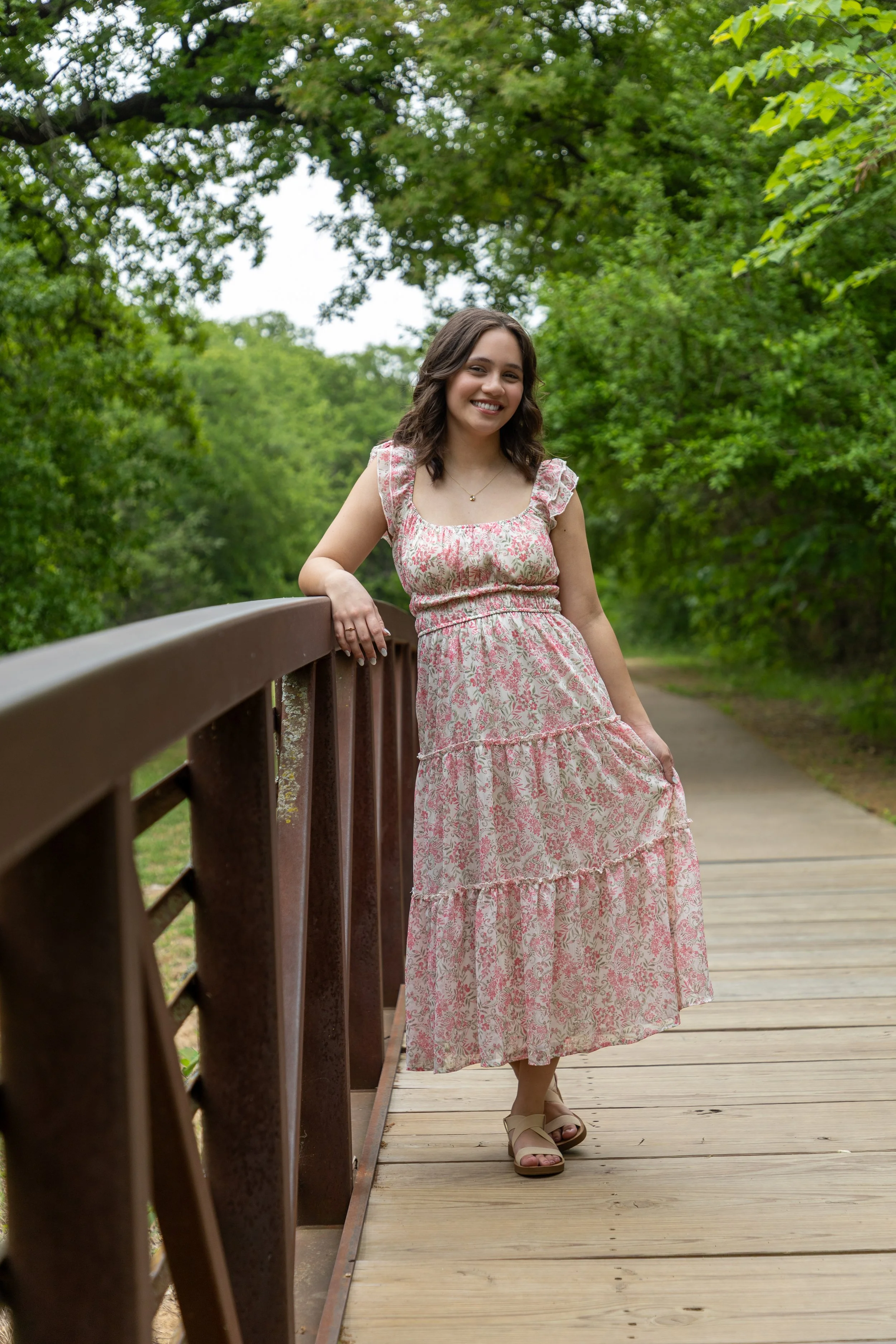 A young woman in a floral summer dress leaning on a wooden railing on a park bridge, surrounded by green trees.