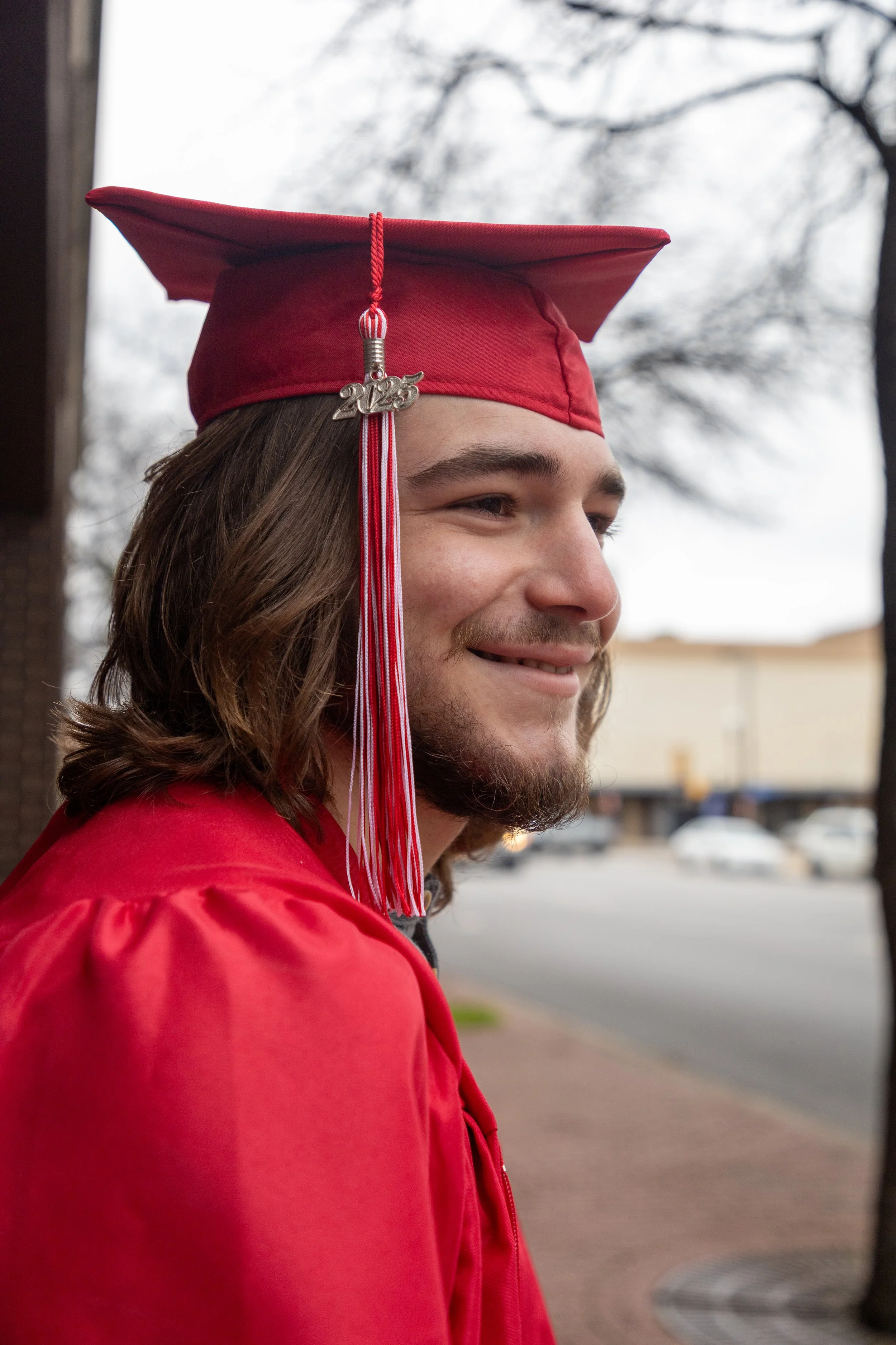 A young man with long brown hair and a beard dressed in a red graduation cap and gown, smiling outdoors during daytime.