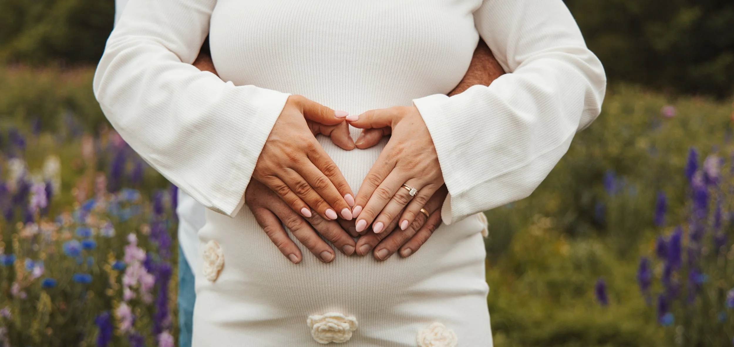 A couple forming a heart shape with their hands over a pregnant woman's belly, standing outdoors in a field of purple flowers.