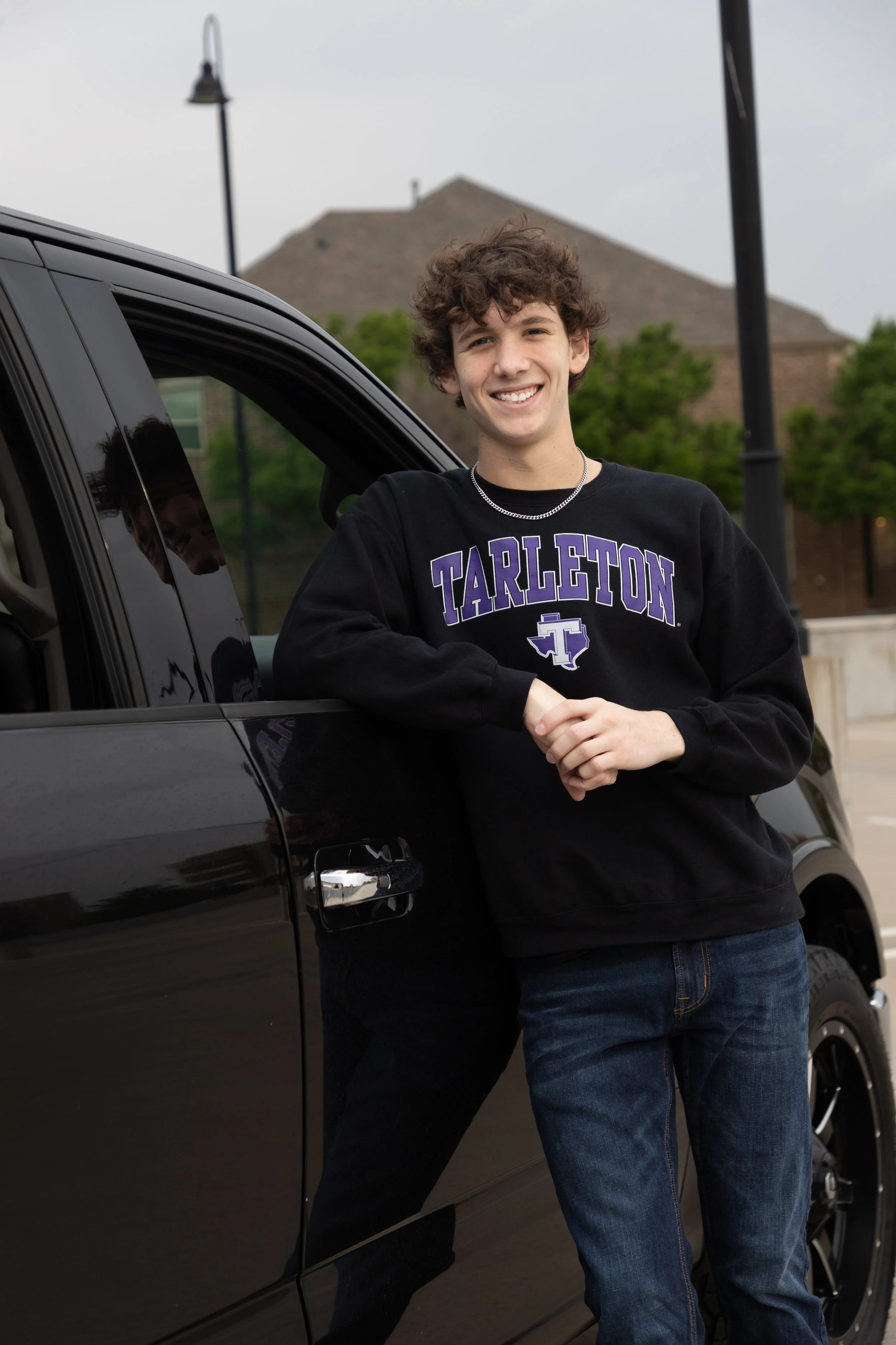 A young man with curly hair smiling and standing next to a black car, wearing a black 'Tarleton' sweatshirt and jeans, outdoors with trees and buildings in the background.