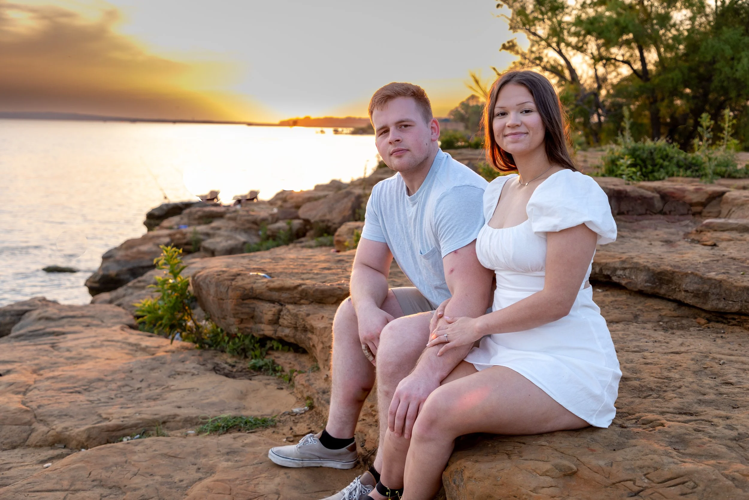 A young man and woman sitting on rocks by a lake at sunset, smiling at the camera.