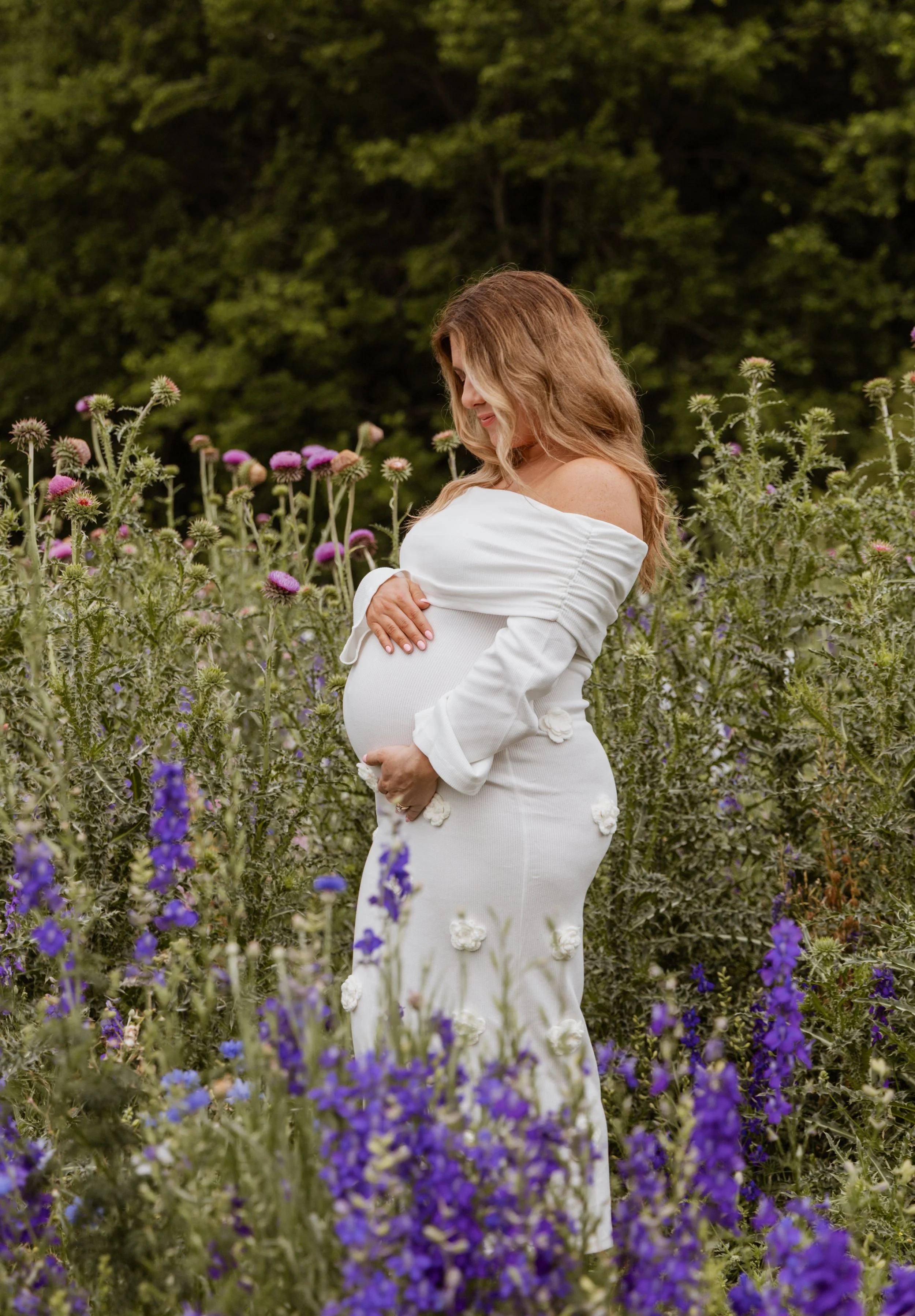 A pregnant woman stands in a field of purple and pink flowers, wearing a white off-the-shoulder dress, smiling softly with her arms crossed over her baby bump, with a background of dense green trees.