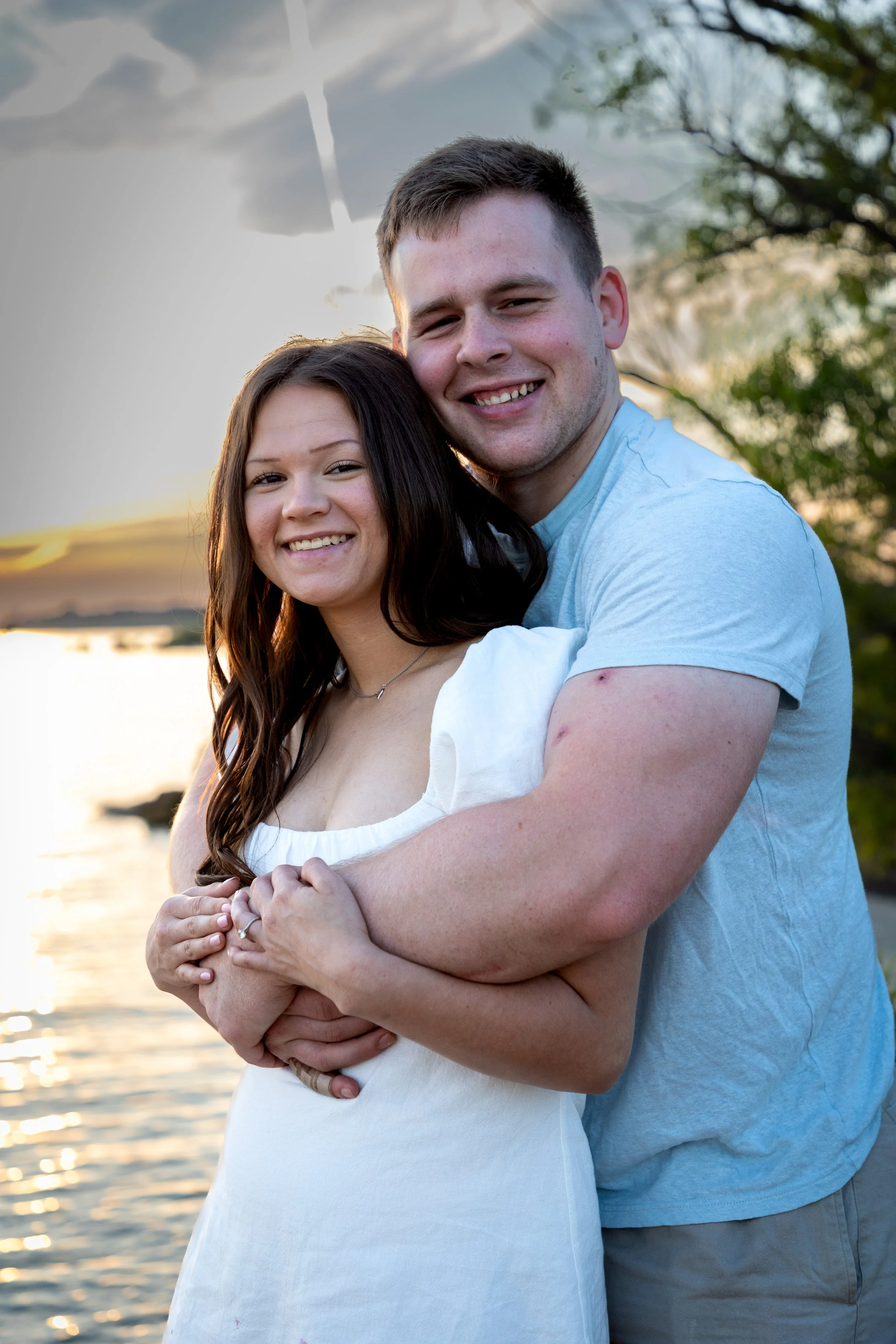 A smiling couple, a woman with long dark hair and a man with short dark hair, embrace each other outdoors near a body of water at sunset, with trees and cloudy sky in the background.