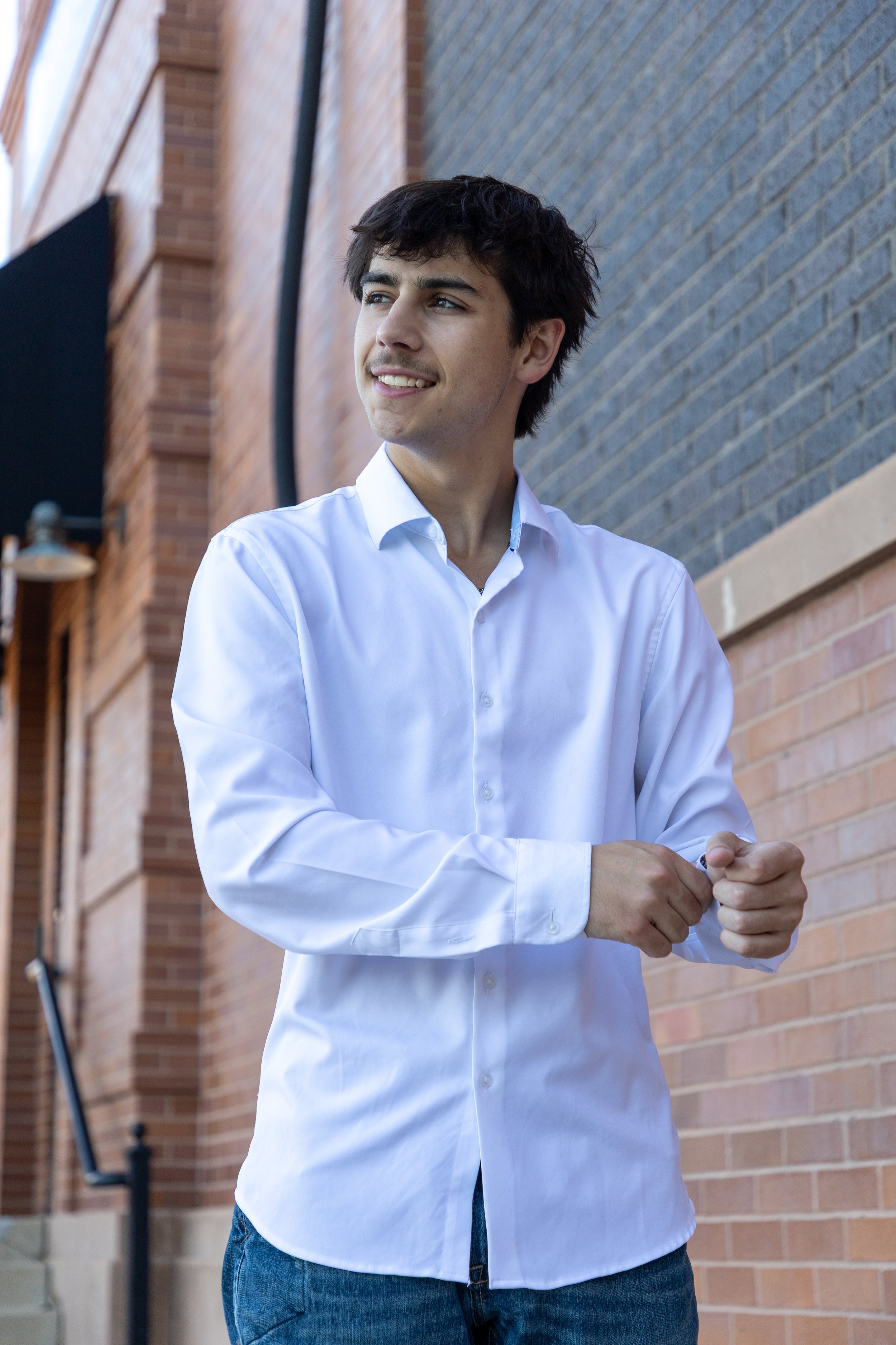 Young man with dark hair and light skin smiling, wearing a white button-down shirt and blue jeans, standing outside against a brick wall, looking to the side.
