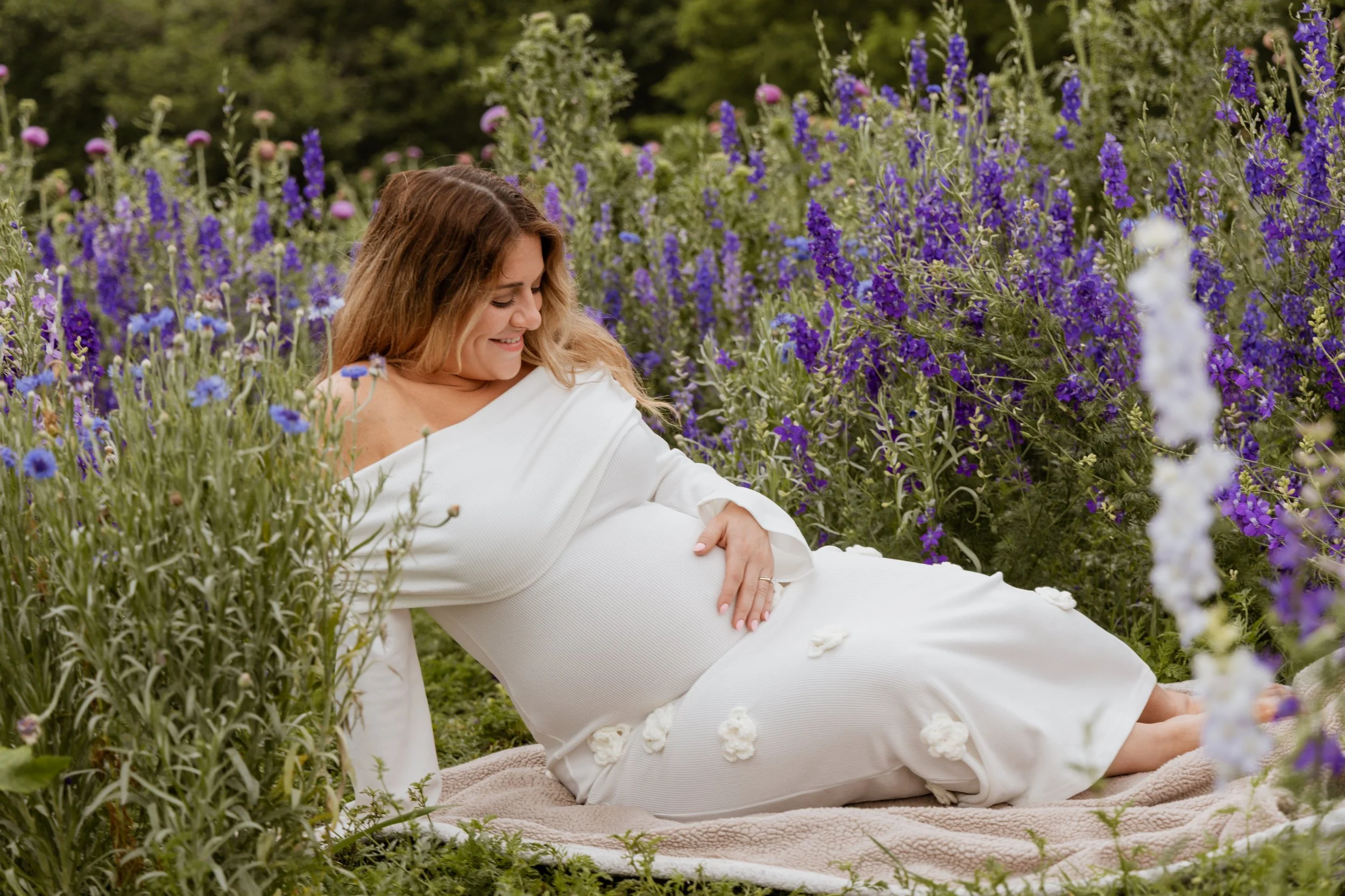 Pregnant woman lying on blanket in a flower garden, wearing white dress and smiling