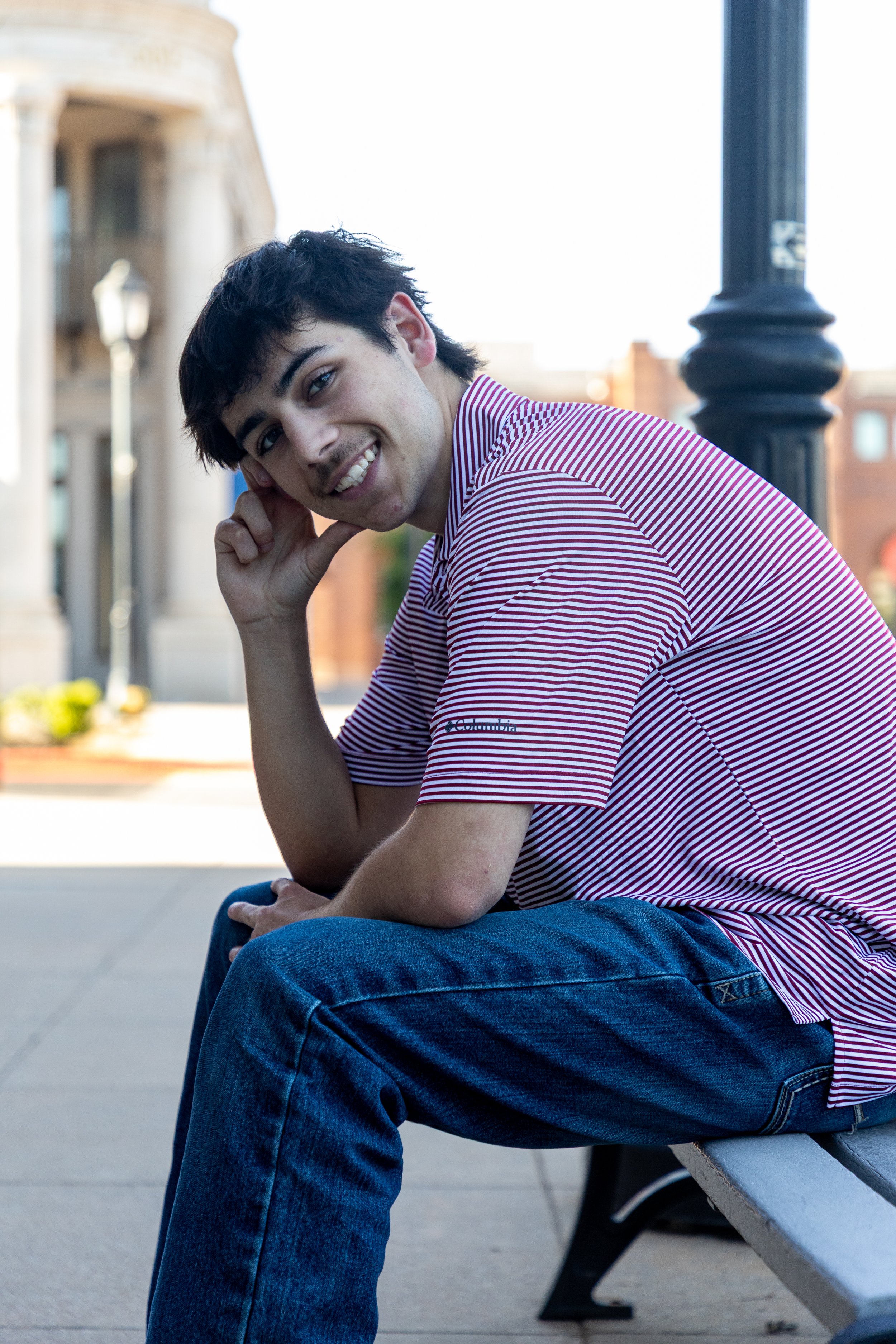 Young man with dark hair smiling and sitting on a park bench outdoors, wearing a striped polo shirt and blue jeans.