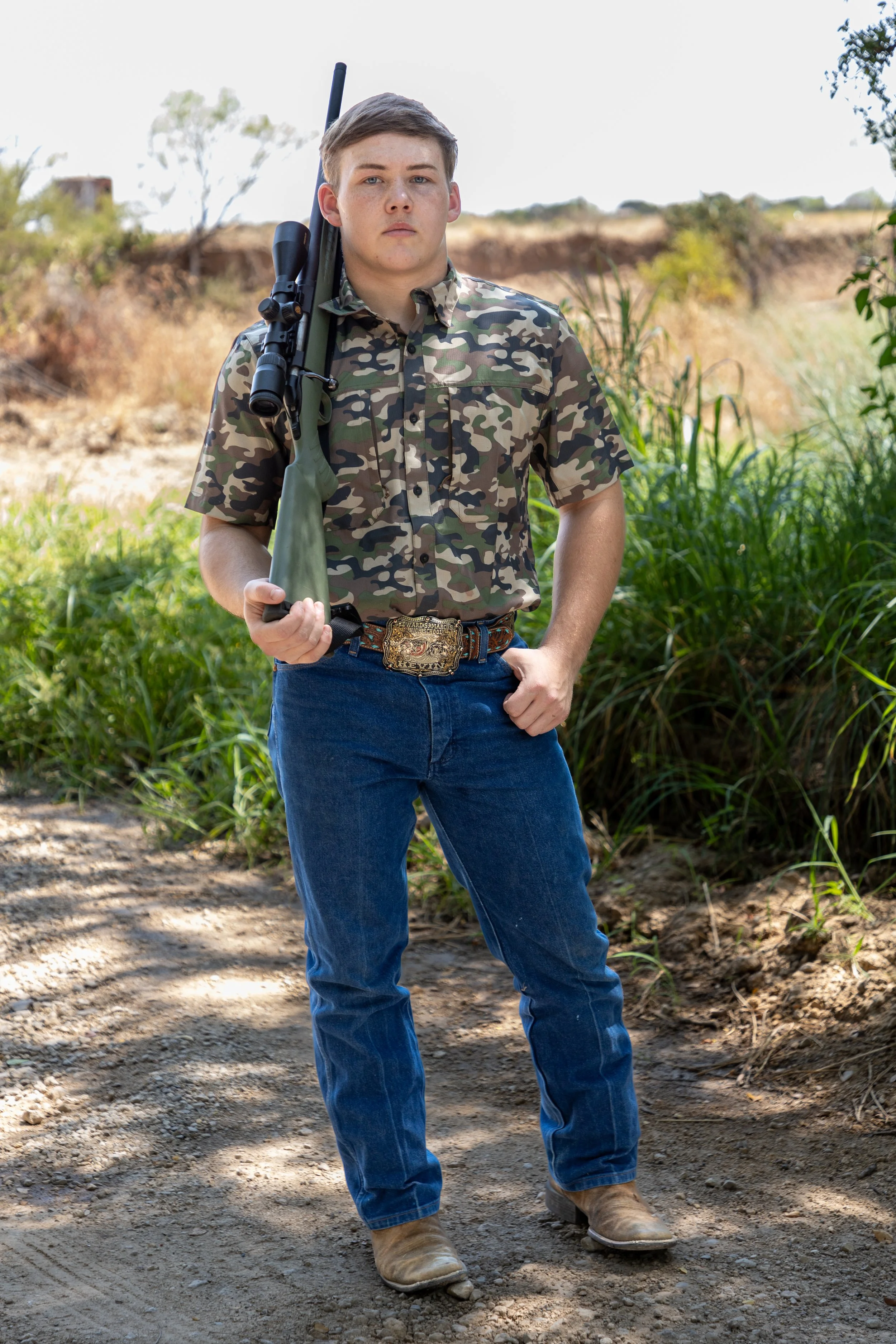 Young man in camouflage shirt and jeans standing outdoors on dirt path, holding rifle, with grassy area and trees in background.