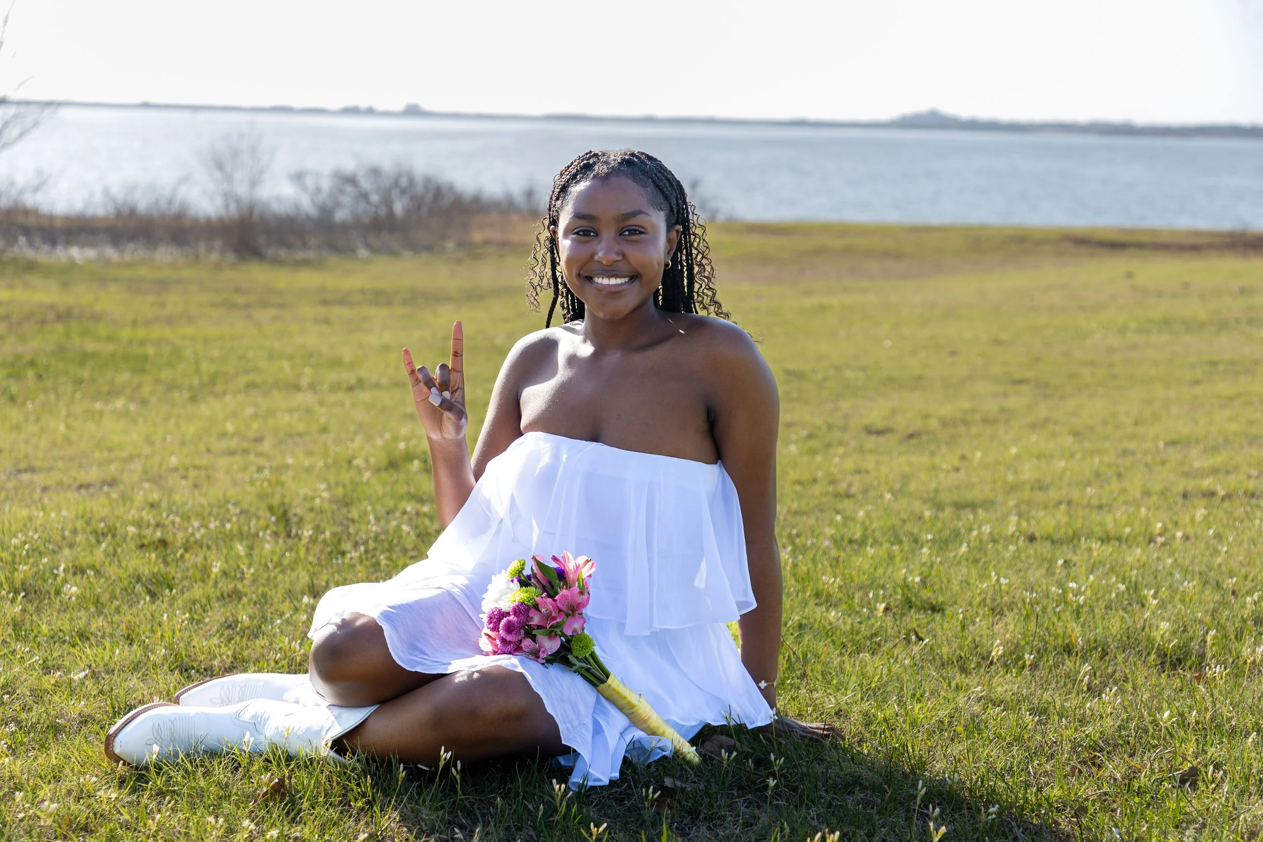 A smiling young woman with curly hair sitting on grass by a body of water, dressed in a white strapless dress, holding a bouquet of pink and white flowers, making a rock gesture with her right hand.