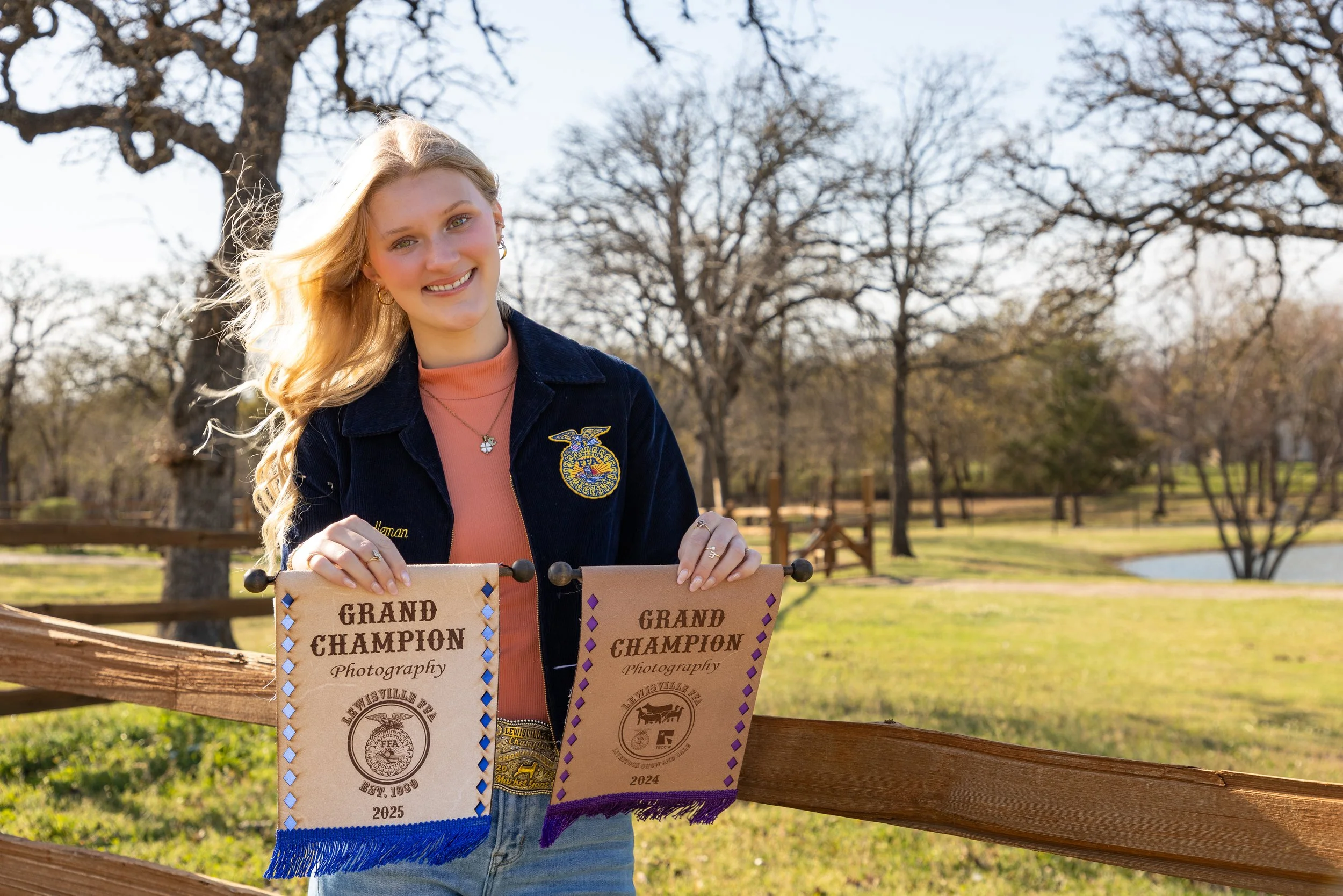 A young woman with long blonde hair holding two 'Grand Champion' banners outdoors in a park with trees and a small body of water in the background.