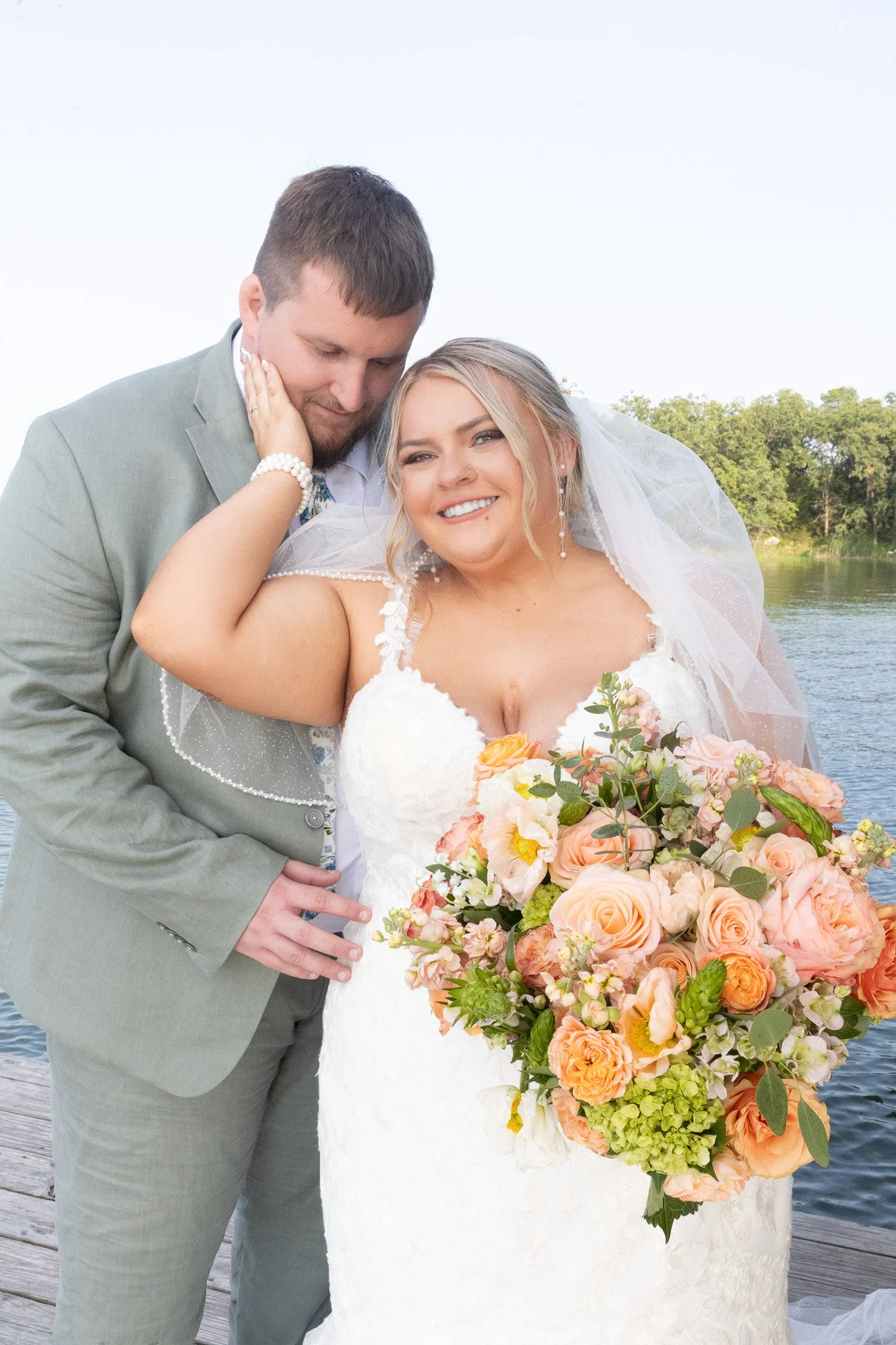 A bride and groom standing on a dock near water, smiling, with the bride holding a large bouquet of peach and white flowers, during a wedding photoshoot outside.