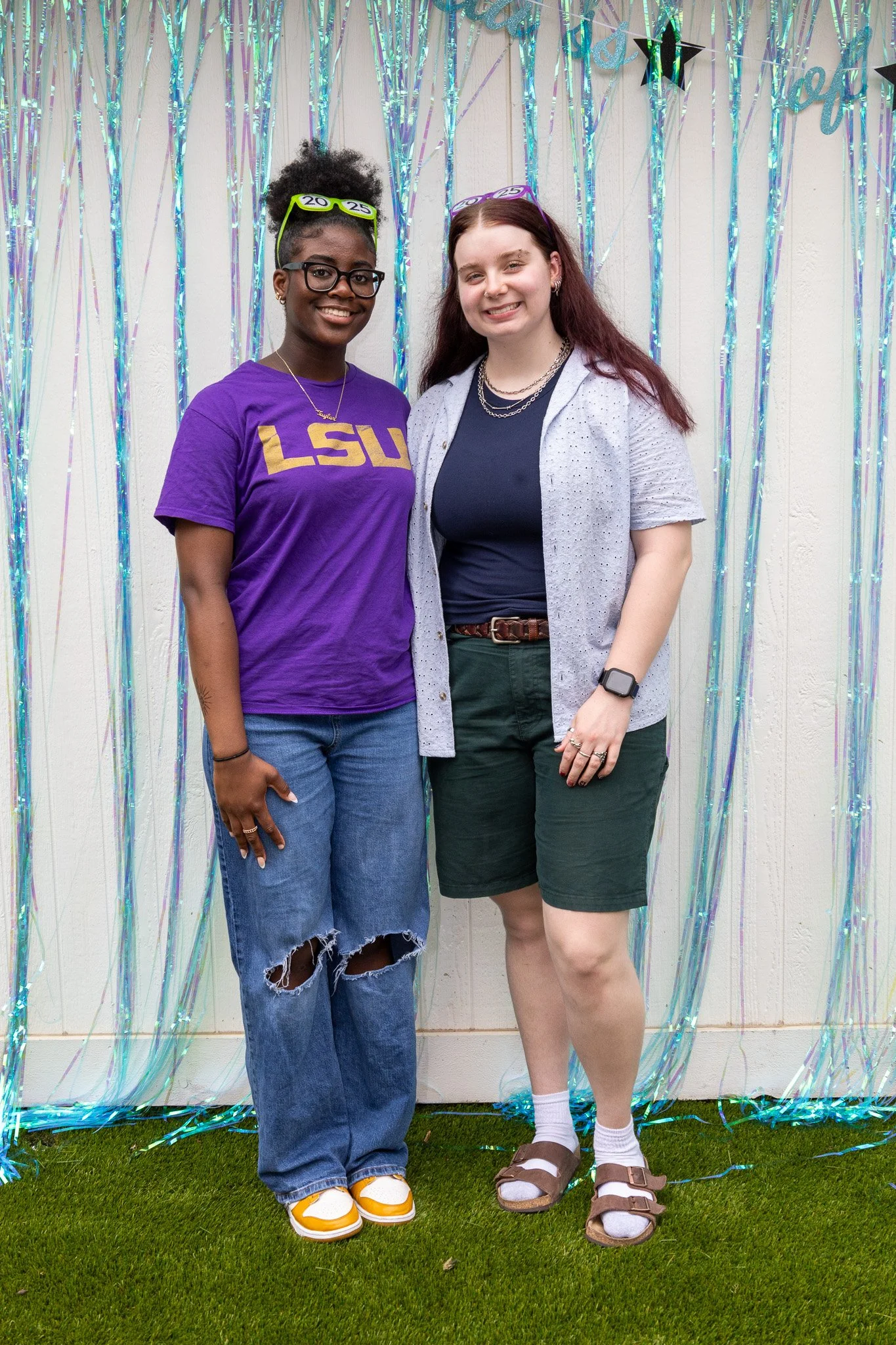 Two women standing in front of a white wall decorated with shiny blue and purple streamers, celebrating a graduation, with one wearing a purple LSU t-shirt and the other wearing a grey shirt and shorts.