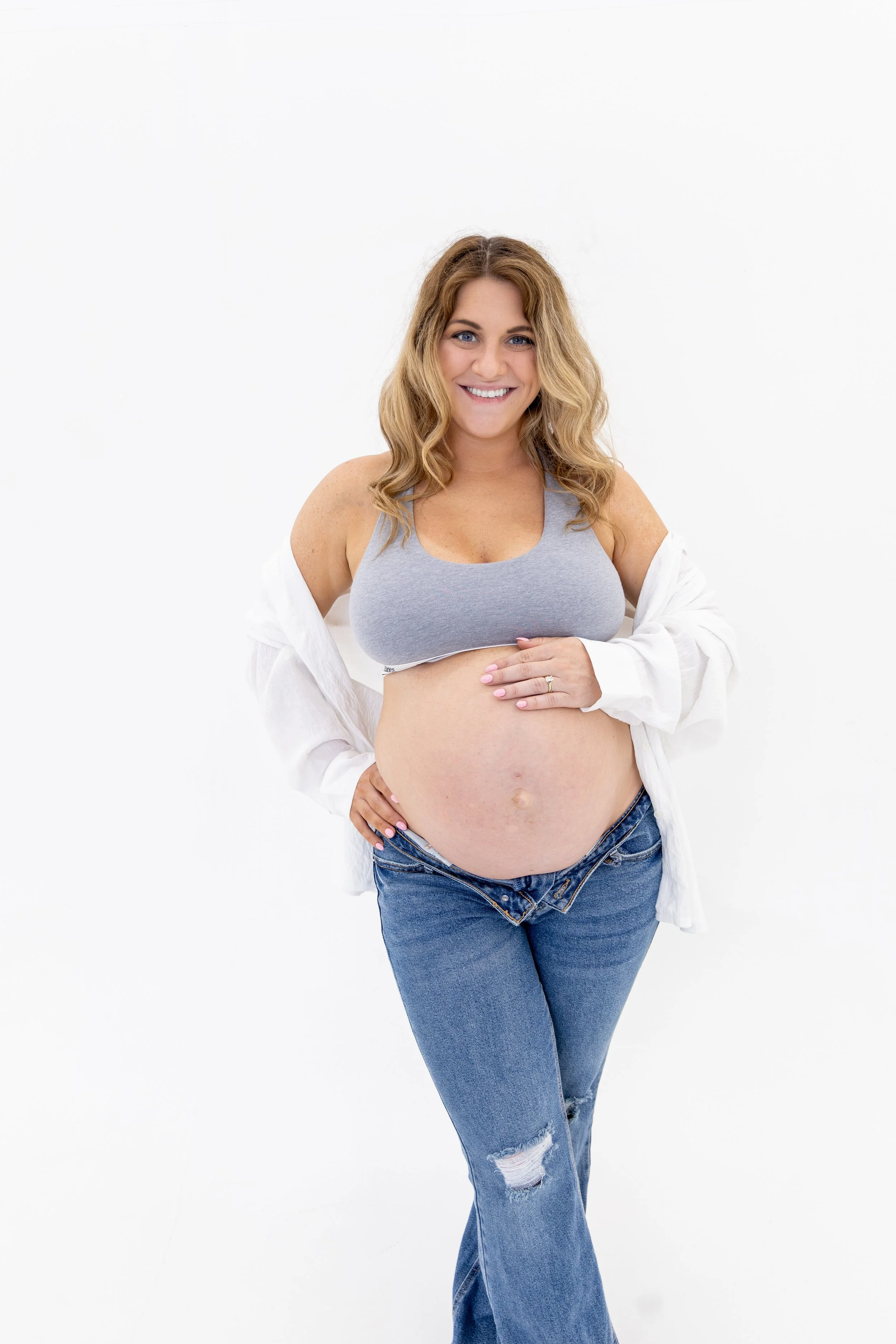 Smiling pregnant woman with wavy blonde hair wearing a gray sports bra, white shirt, and blue jeans with a rip in the knee, standing against a plain white background.