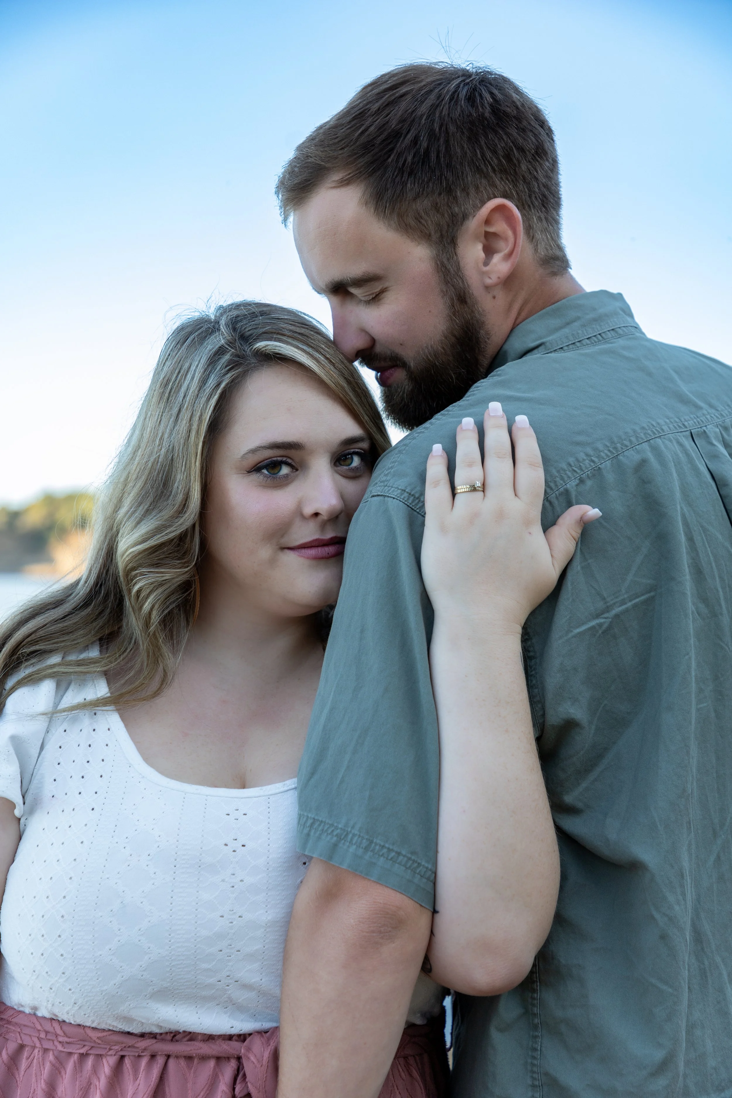 A young woman with long blonde hair and a man with a beard embrace outdoors, with the woman resting her hand on the man's shoulder, displaying a wedding ring.
